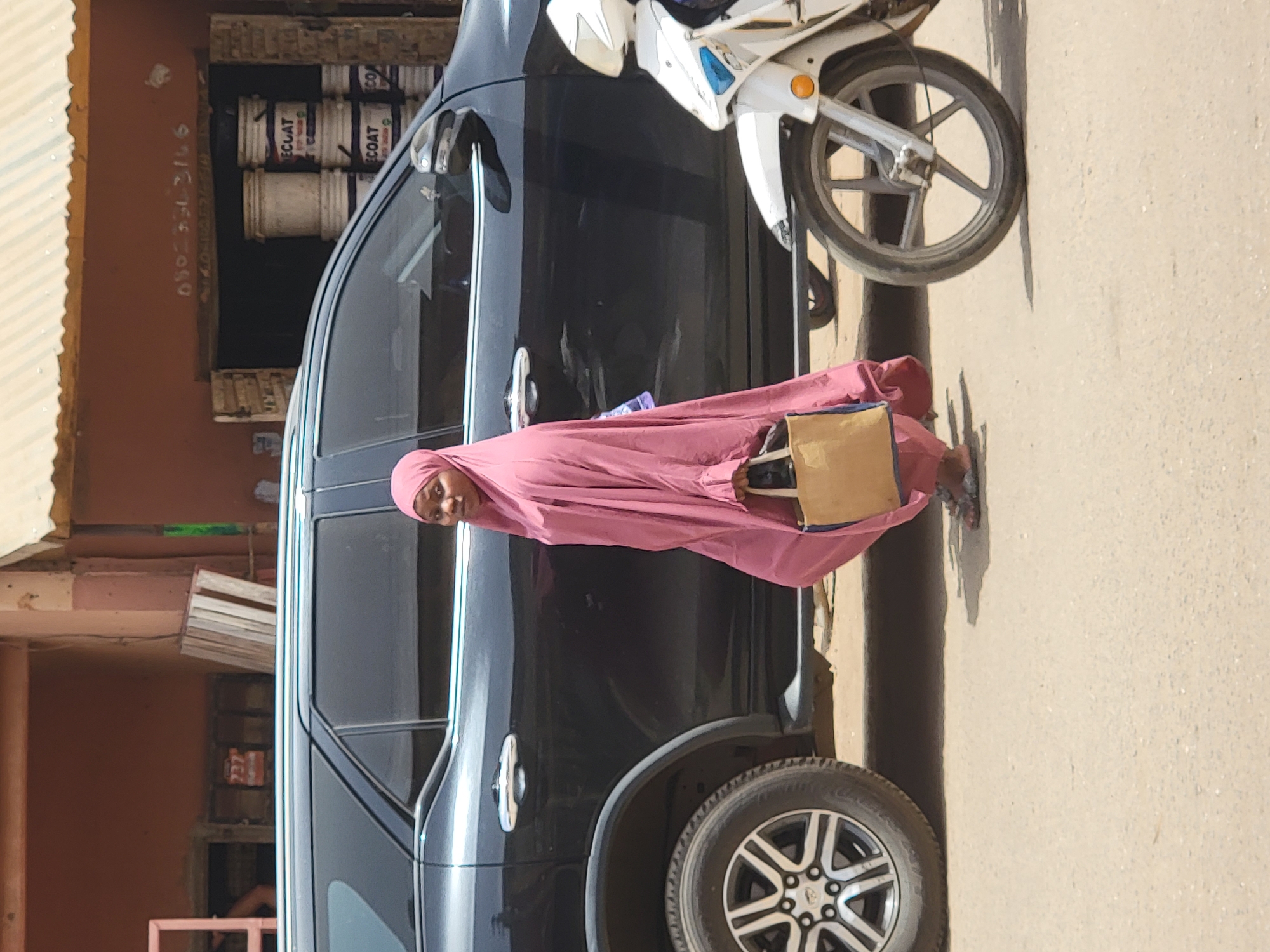 An outdoor scene depicts a woman standing on a dry, dusty street in Maiburiji, Nigeria, under bright daylight. The woman, possibly young, is centrally positioned, dressed in a light pink head covering and a loose, full-body pink garment. She holds a light brown bag and looks directly at the viewer with a gentle smile.
To her left, a black SUV is parked, its side reflecting the bright sun. Behind the SUV and slightly to the right, a white scooter or motorcycle is also parked. The ground beneath the vehicles and the woman is light-colored and appears dusty, with visible shadows cast, suggesting a sunny day, likely midday.
In the background, to the left of the SUV, an earthy-toned building wall is visible, partially obscured by a corrugated metal structure. An opening in the wall reveals stacked white containers, some clearly labeled "ECOAT," indicating a possible paint or hardware store. On this wall, a handwritten phone number "080-7313106" is visible.
The overall atmosphere is casual and everyday, capturing a moment on a street in what appears to be an urban or semi-urban setting.