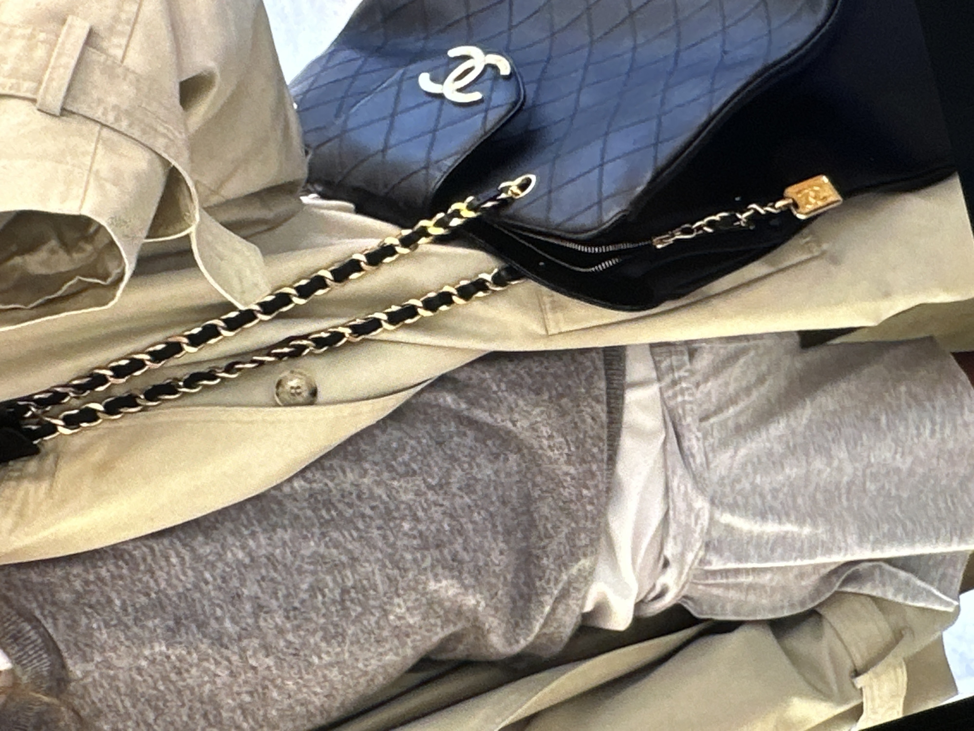 A close-up shot presents a collection of clothing and accessories. A navy blue Chanel bag with a quilted pattern and intertwined C logo rests on a tan trench coat with a button detail. A gray sweatshirt and a white garment are also present. The scene seems to be indoors. Information about the city of Yerevan, Armenia, is not evident in the picture.