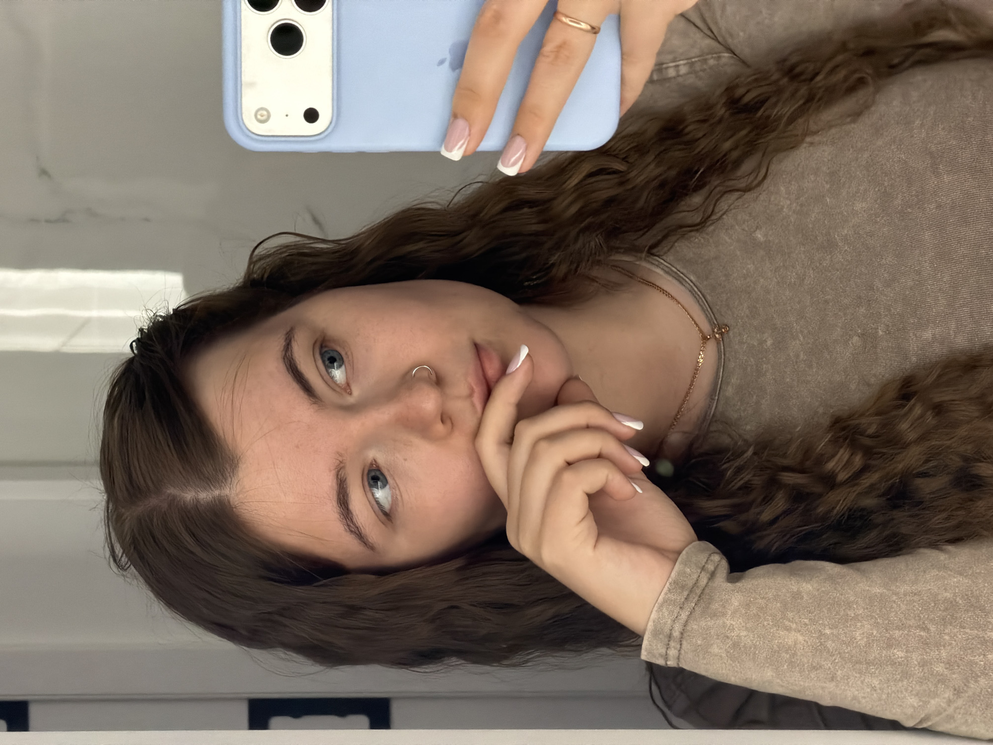 The image shows a young woman taking a selfie in what appears to be a bathroom. She has long, curly brown hair, blue eyes, and a nose ring. She is wearing a tan shirt and a delicate gold necklace. Her nails are painted with a white tip. She is holding a light blue smartphone up to take the photo.
There is no information in the image or given context to suggest this image was taken in the city of Chaikovsky, Russia. The surrounding environment appears to be a bathroom with a white wall and a visible reflection in a mirror.