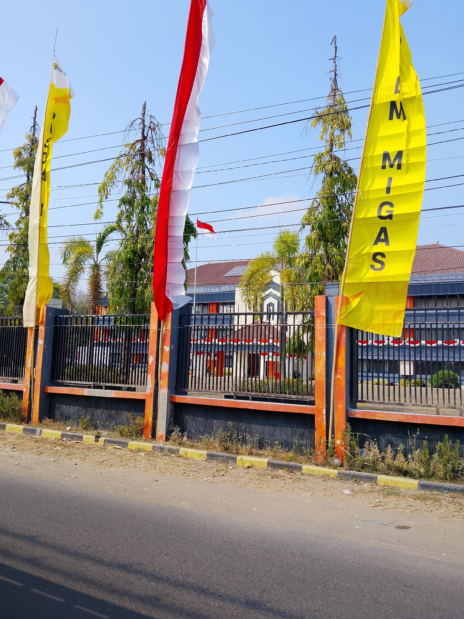 The image shows a street scene in Ngelo, Indonesia. In the foreground, a paved road runs horizontally across the bottom of the frame, with a dirt shoulder. A black metal fence with orange and gray posts runs along the side of the road, separating it from a building in the background. Several flags are visible, including a tall yellow flag with black lettering that appears to say "IMIGAS", and a red and white flag, which is the national flag of Indonesia. The building behind the fence is a two-story structure with a tiled roof and windows. There are trees and some vegetation growing near the fence. The sky is clear and blue.