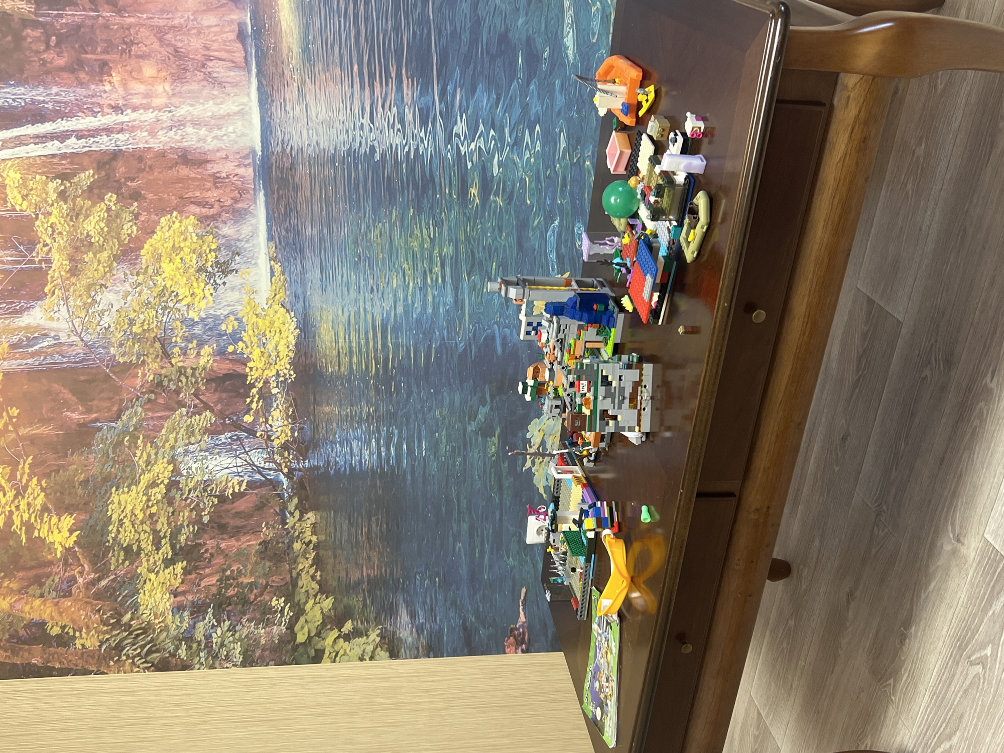 The image shows a wooden coffee table with various LEGO builds on its surface. The background features a large mural depicting a waterfall and a body of water, possibly in a natural setting. On the table, there are several distinct LEGO constructions, including what appears to be a small building with a bicycle on top, a boat with a crane, and various smaller structures and individual pieces. A green LEGO brick is visible on the table surface. The table itself is dark wood with two drawers. The floor is made of light-colored wood planks. There is no indication of the city of Volgograd, Russia in the image.