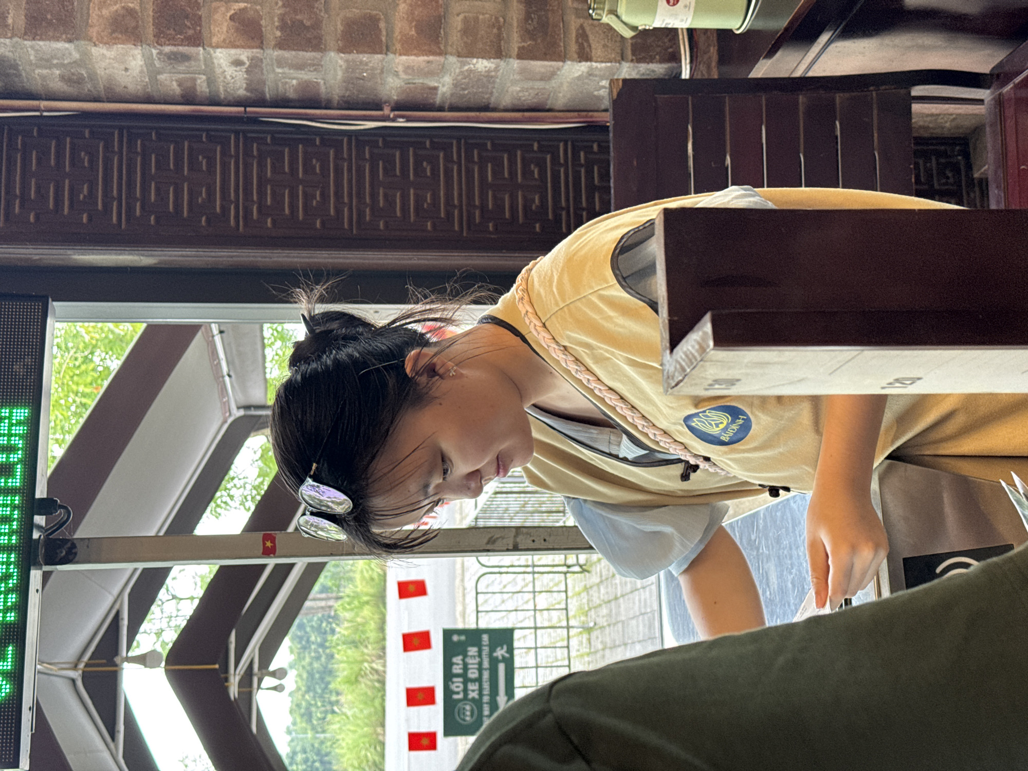 In Tây Hoa Lư Ward, Vietnam, a young East Asian woman with her hair tied up and sunglasses on her head is seen from the shoulders up. She is wearing a light yellow vest over a light blue shirt. She is holding a white ticket or card in her hands, and appears to be interacting with someone out of frame to her left. Behind her, a large green digital sign is visible, and in the background, there are red flags and trees. The environment appears to be an indoor/outdoor setting, possibly a ticket booth or an entrance area, with brick walls and dark wooden decorative elements. A green water bottle is visible to the right.
