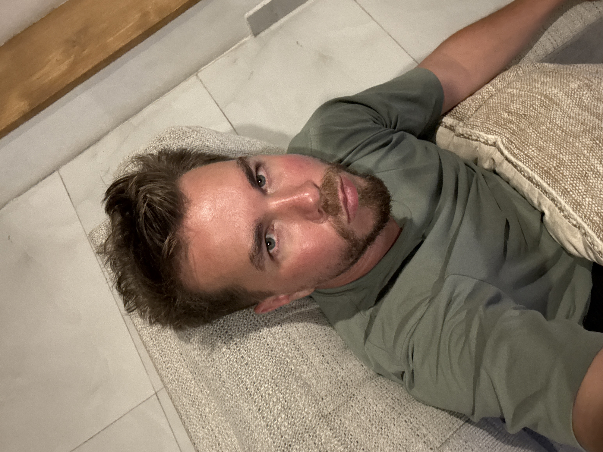 A Caucasian man is lying on a textured surface resembling a couch or large cushion in what appears to be an interior setting. The man is fair-skinned with brown hair and a short beard. He is wearing a green short-sleeved shirt. He is looking directly at the camera. He has a tan-colored pillow behind his head. The floor is tiled. The lighting appears to be indoor lighting. The scene may be located in North Kuta, Indonesia.