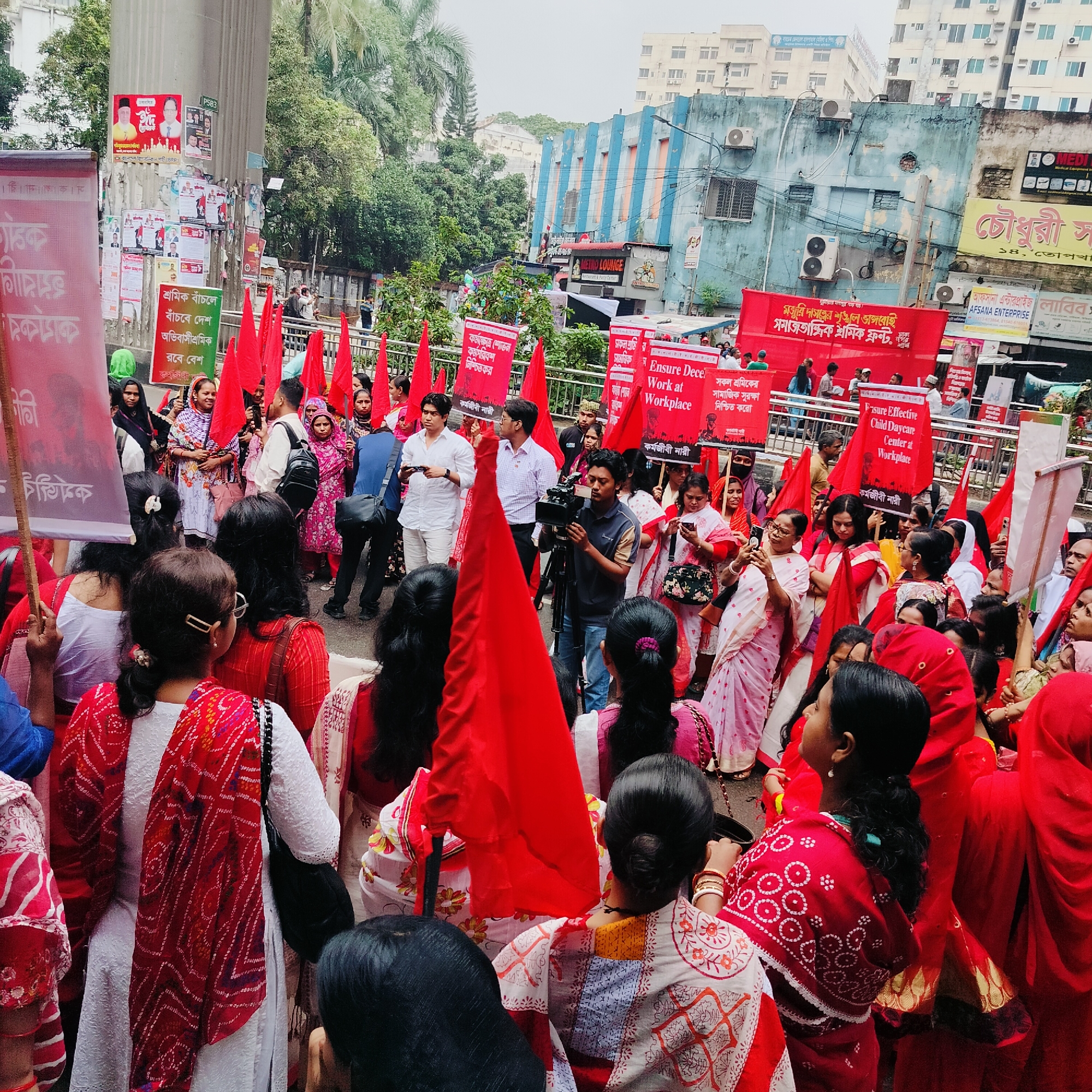 A crowd of people, predominantly women, is gathered in what appears to be a street protest or demonstration. Many are carrying red flags and signs with text written in Bengali. The crowd is dense, with people wearing a mix of traditional and modern clothing. The color red is prominent in the attire of many protesters, including red sarees, scarves, and banners. A cameraman with a professional video camera is recording the event.
The background includes buildings with signs written in Bengali, indicating the location is likely in Bangladesh or West Bengal, India. A building labeled "METRO LOUNGE Restaurant & Fast Food Center" can be seen. Other businesses and signs are visible in the urban environment. There is a concrete structure or column with posters and notices affixed to it. The weather appears overcast.