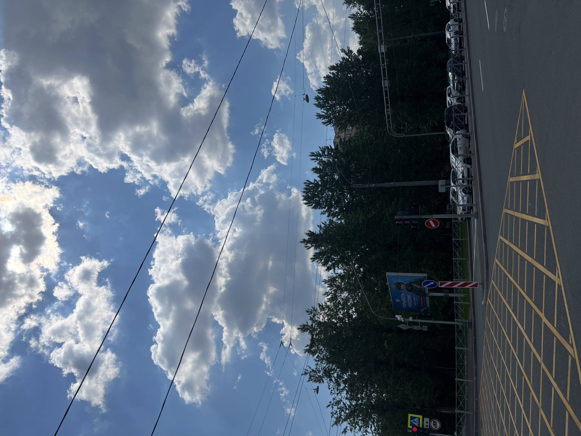 The image captures a vibrant daytime urban street scene in Saint Petersburg, Russia. The sky dominates the upper portion, a bright blue canvas dotted with numerous fluffy white cumulus clouds, suggesting clear and sunny weather. Several dark utility lines stretch across the sky, supported by unseen poles, intersecting the cloud formations.
On the right side of the image, a paved asphalt road is visible. It features distinct yellow grid markings, characteristic of a box junction or a no-stopping zone, alongside standard white lane dividers. To the left of the road, a row of diverse cars, predominantly white and dark, are parked in an orderly fashion behind a protective metal barrier. A dense line of green trees provides a natural border behind the parked vehicles.
Several urban infrastructure elements are present: at the bottom left, a traffic light displays red and yellow signals, with a red circular 'no entry' sign visible above it. Further along the street, a blue billboard stands prominently, featuring an individual in military uniform. The billboard displays Cyrillic text that reads "ГОРДОСТЬ РОССИИ!" (PRIDE OF RUSSIA!), confirming the Russian context. Below this, a blue circular sign with a white arrow points to the right. The scene is devoid of visible people, focusing instead on the city's infrastructure and natural elements under a bright sky. The overall impression is one of a typical, active urban environment during daylight hours.