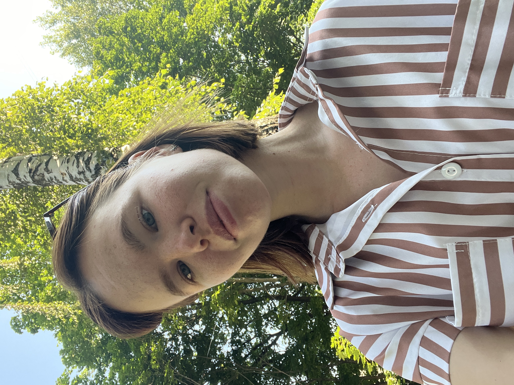 A young woman with short brown hair and freckles is taking a selfie outdoors. She is wearing a brown and white striped shirt. Behind her, lush green trees and a blue sky are visible. The image appears to have been taken on a sunny day. The location is Chaikovsky, Russia, specifically in a park or wooded area, given the natural surroundings.