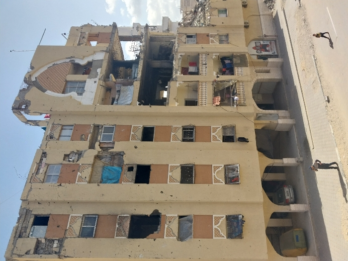 An elevated view captures a multi-story residential building displaying extensive structural damage in Khan Yunis, Palestinian Territory, under a clear sky. The building's light-colored facade is heavily scarred with numerous impact points consistent with bullet or shrapnel damage. The right side of the structure has suffered catastrophic damage, with significant portions of upper floors and external walls completely obliterated, exposing internal rooms, rebar, and debris. A distinctive arched architectural element at the building's apex on the right is partially destroyed.
Many windows across the facade are broken, missing, or obscured by makeshift coverings, including blue tarpaulins and dark sheeting. Laundry is visibly hanging from several balconies and within exposed window frames on the lower-mid levels. The ground floor incorporates a series of arched openings leading to a covered area, where a yellow-green vehicle covered by a tarp and a silver sedan are parked.
On the street level below, an adult male in dark clothing and a cap stands on a dirt shoulder next to a paved road, appearing to hold a phone to his ear. Further to the right, a young child walks or runs on the paved road. Both individuals cast distinct shadows to their right. A red and white poster with text and an image is affixed to the lower right section of the building's facade.