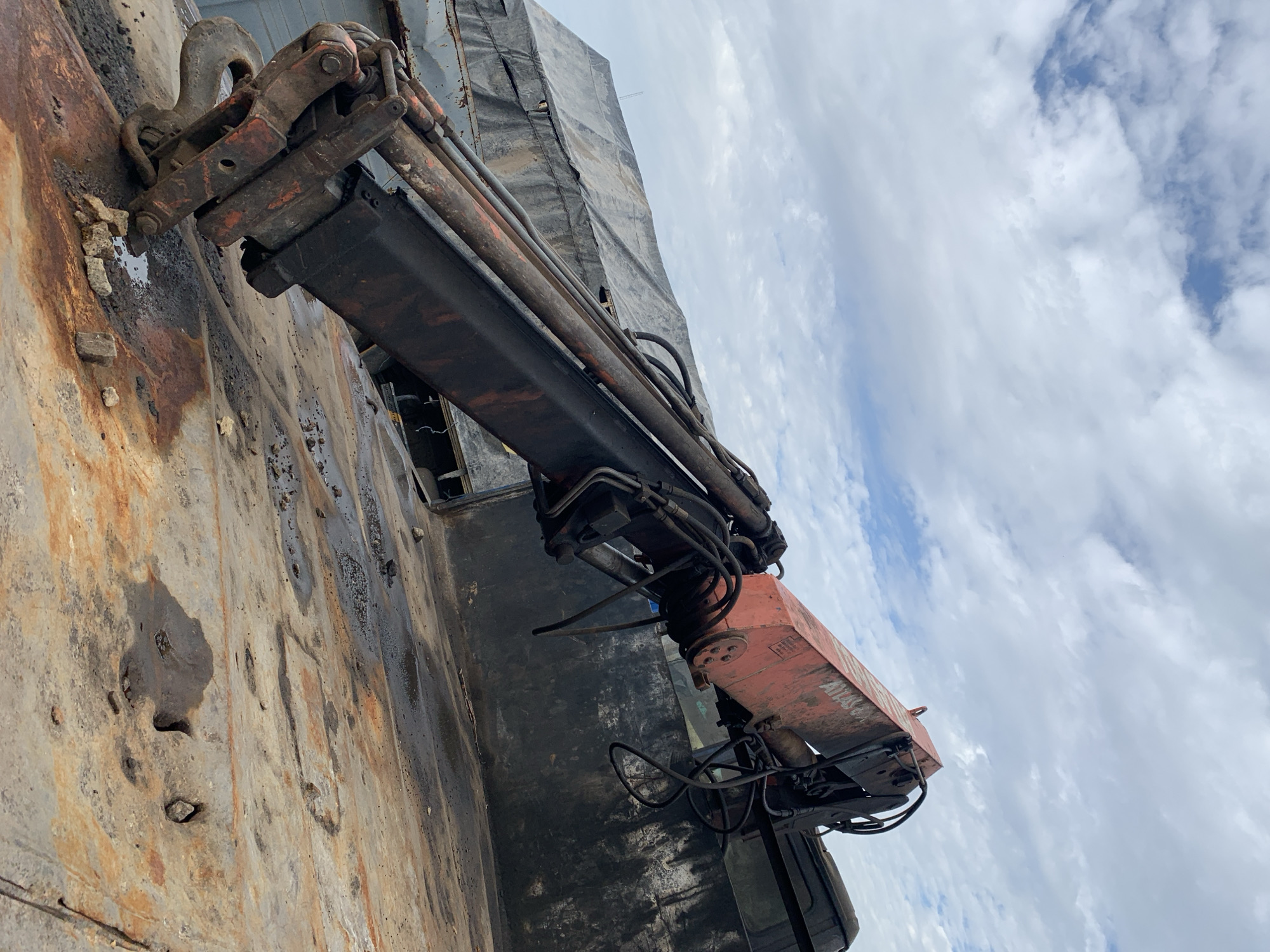The image captures a static, heavily weathered red and black ATLAS hydraulic crane arm, identified by partial "ATLAS" lettering on its upper section, positioned on a corroded metal deck under an overcast sky. The crane's robust hook rests on the rusty deck, which shows extensive orange and brown discoloration, dark fluid stains, and scattered small rocks and debris. In the background to the left, a large structure draped with a grey tarpaulin is visible, while to the right, a dark, grimy metallic wall or surface is present. The scene is an outdoor, industrial environment, likely a port or shipyard, located in Rafin Par, Nigeria. No individuals or active operations are depicted.