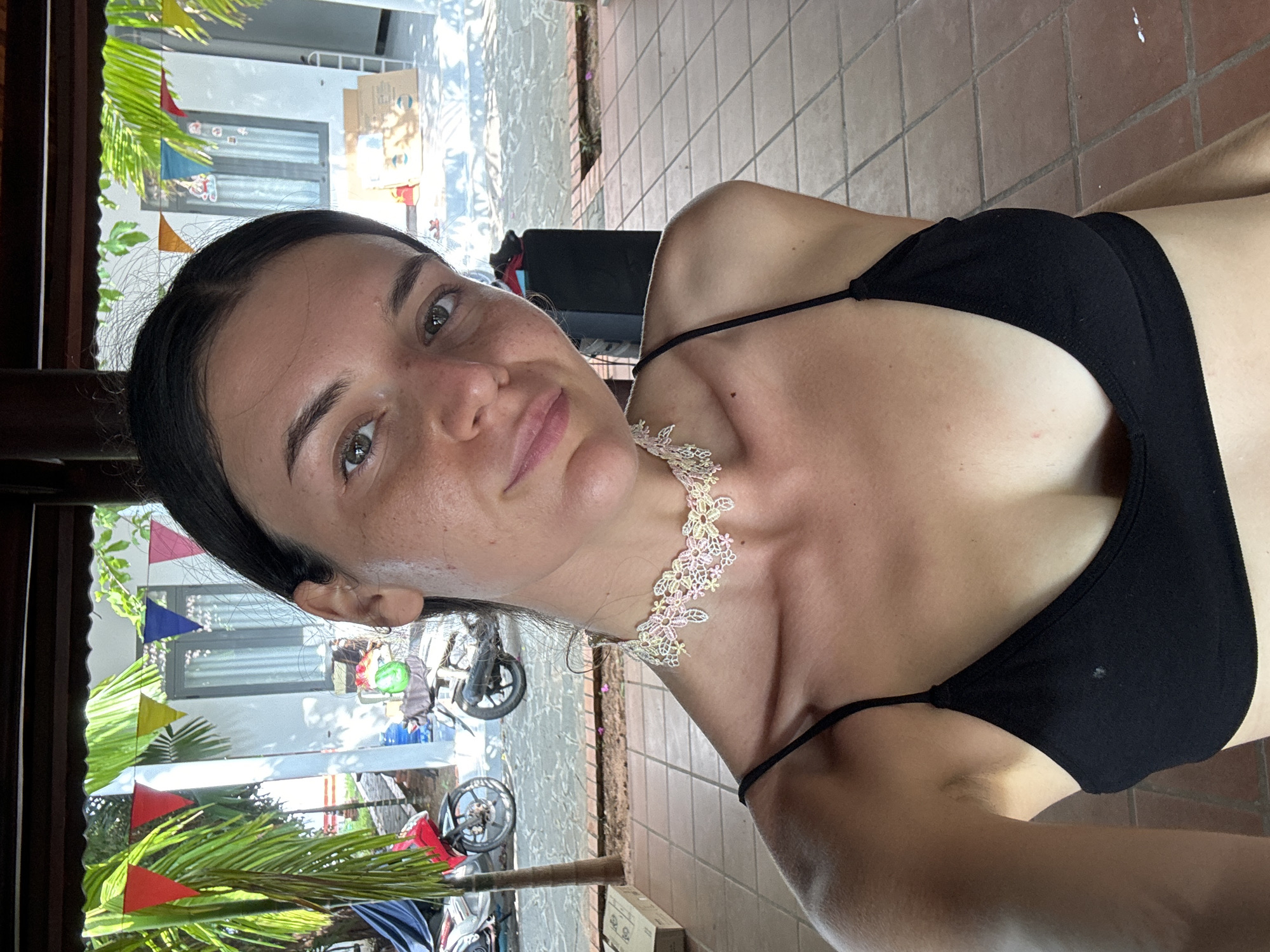 A young woman with dark hair and green eyes, wearing a black tank top and a delicate lace choker, smiles at the camera. She is positioned in the foreground, with the background showing a covered outdoor area with tiled flooring. Behind her, a white building with windows is visible, adorned with colorful triangular flags. Several motorcycles are parked outside, and palm trees with green fronds can be seen to the left. The scene is illuminated by natural sunlight, casting shadows on the ground. The overall impression is that of a casual, sunny day in a tropical location. This image was likely taken in Mũi Né, Vietnam, known for its beaches and coastal resorts.