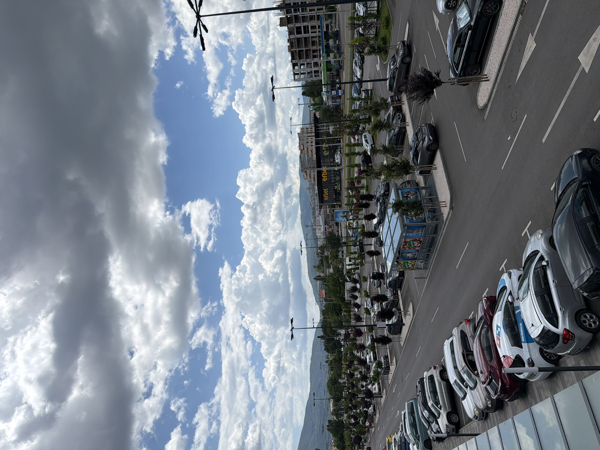 The image depicts an outdoor parking lot in Sofia, Bulgaria, under a cloudy sky with patches of blue. The parking lot is filled with numerous cars of various makes and models. In the background, there are several buildings, including a multi-story structure under construction and a commercial building with signage that appears to read "eFbet". A gas station is also visible. Trees are planted in rows throughout the parking lot, and lampposts are strategically placed for illumination. The scene appears to be a typical urban environment, likely near a commercial area or shopping center, on a day with dynamic weather conditions. No people are clearly visible in the frame.