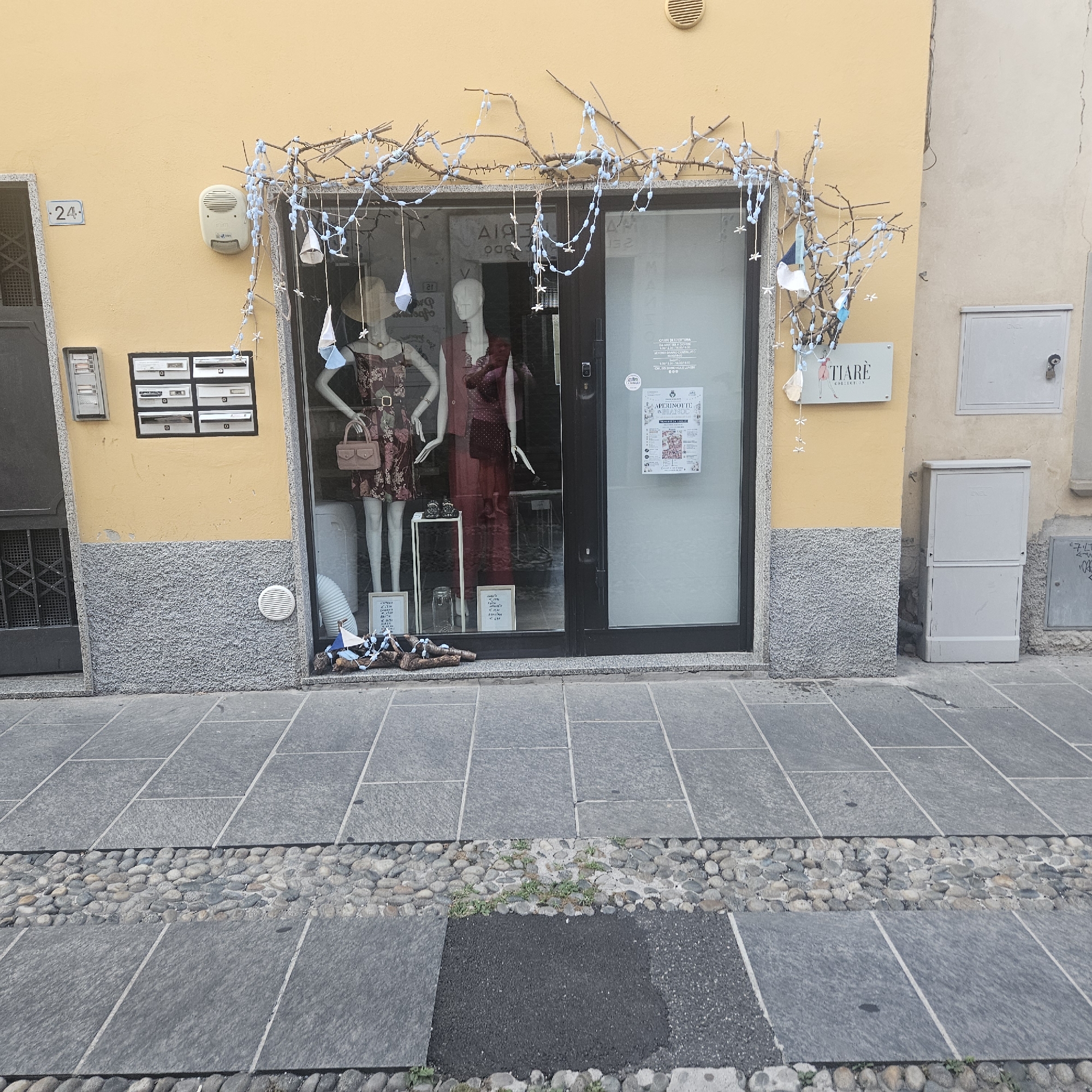 The image shows the facade of a shop called "TIARÈ COLLECTION" in Calcinate, Italy. The shop has a large display window showcasing two mannequins dressed in women's clothing. One mannequin on the left wears a floral print dress with a belt and carries a small pink handbag. The other mannequin on the right is dressed in a burgundy-colored dress and a matching blazer. Both mannequins have their heads covered with straw hats. The shop window is decorated with a string of blue lights and delicate hanging ornaments resembling small boats and flags. Inside the display, there are framed pictures and a glass jar. The exterior wall of the building is painted yellow, with a textured grey section below the window. To the left of the shop entrance, there is a gray metal door and a set of mailboxes. Above the mailboxes, a white intercom or alarm device is mounted on the yellow wall. The number "24" is visible on a small blue plaque on the wall. The pavement in front of the shop is made of large grey stone slabs interspersed with a band of small grey and brown pebbles. A light grey electrical box is mounted on the right side of the building, next to the shop entrance.