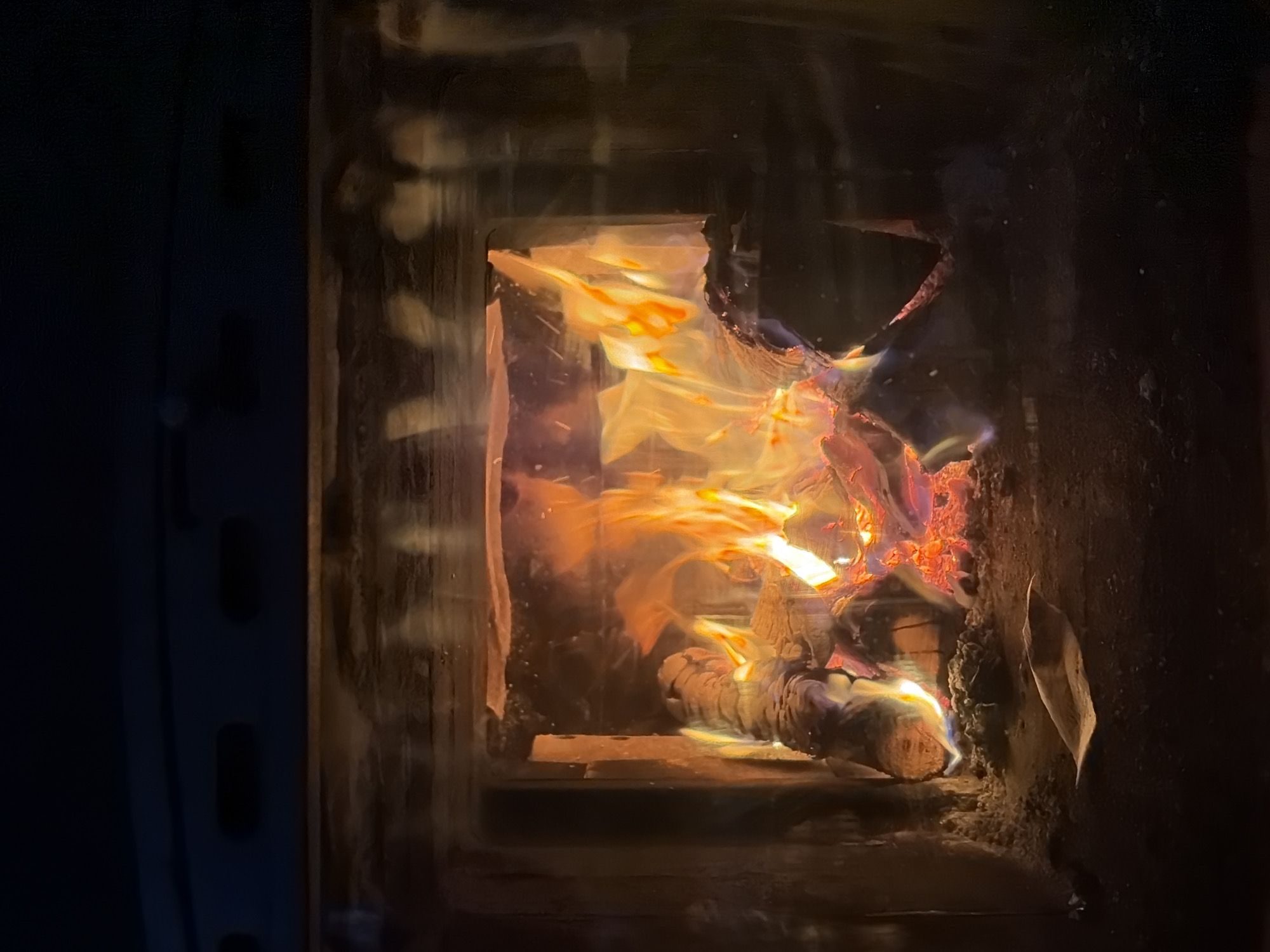 A close-up view of a wood-burning stove with flames emanating from burning wood inside. The stove is located in an interior space, and the foreground consists of the dark metallic structure of the stove. There are visible flames and burning embers inside the stove. The location is Кадряково, Russia.