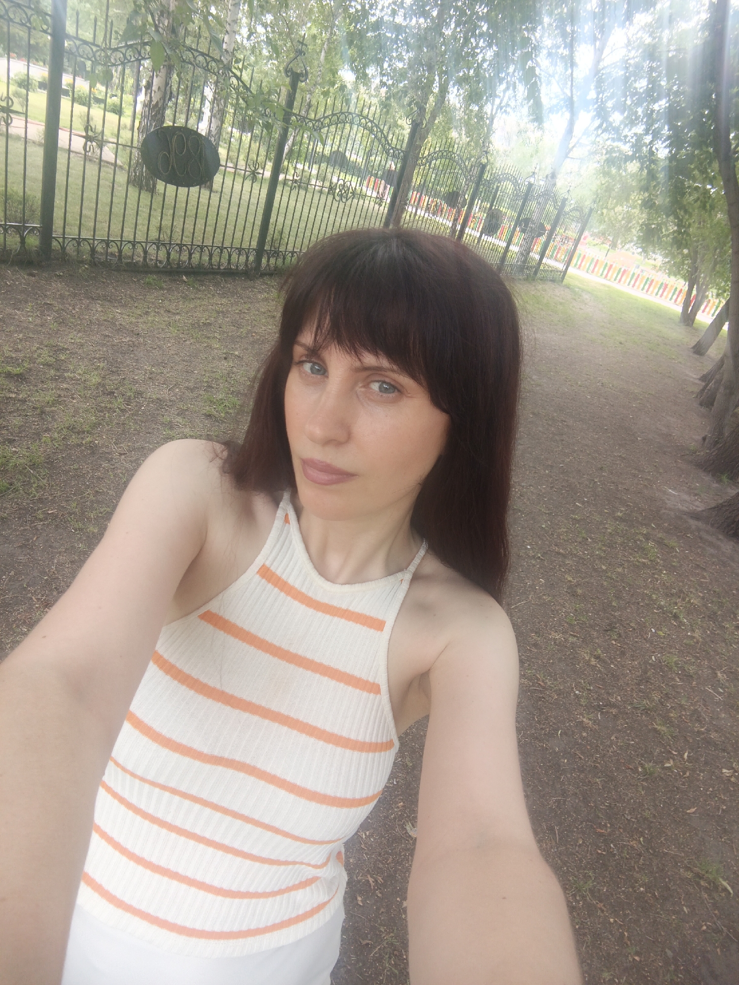 A woman with brown hair and bangs, wearing a white and orange striped halter top and white shorts, takes a selfie outdoors. She is positioned on the left side of the frame, with her body angled towards the right. Behind her, a black wrought-iron fence encloses a grassy area with trees. The ground is a mixture of dirt and dry grass, with a path to the right lined with trees. The sky is visible through the foliage. The scene appears to be a park or public space in the city of Kurgan, Russia.