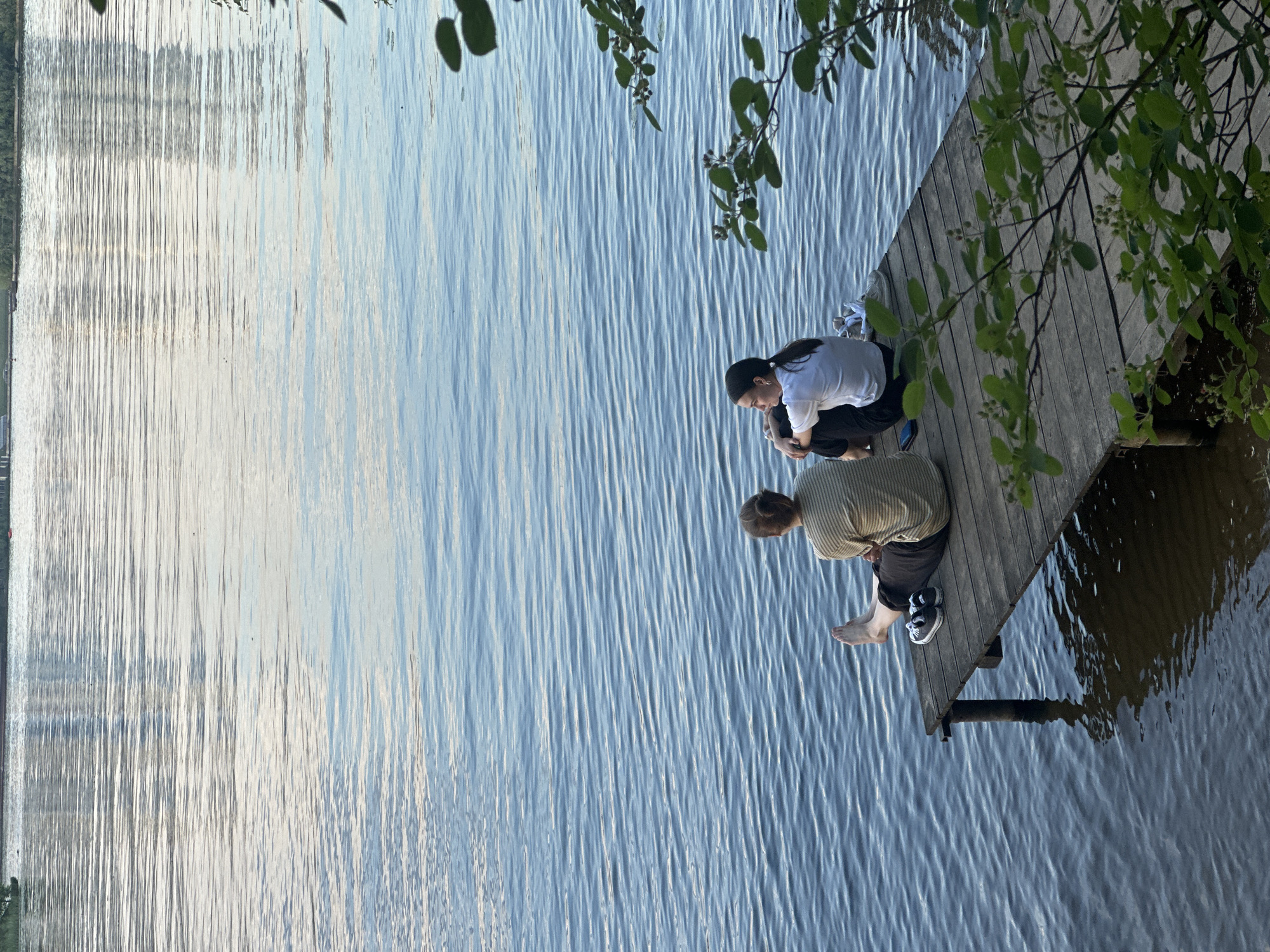 Two individuals are seated on a rustic wooden dock, extending into a calm body of water, likely a lake or wide river. The scene is dominated by the expansive water surface, which reflects light, creating shimmering, elongated patterns, especially on the left side of the frame.
The person closer to the viewer, positioned on the left side of the dock, wears a light-colored, possibly striped, short-sleeved top and dark trousers. One foot is bare, while the other sports a black and white sneaker, suggesting a moment of relaxation or contemplation. Their hair is light-colored and short. They are looking down towards the water.
Further along the dock, to the right, sits a second person wearing a white t-shirt and dark pants. They have dark hair, tied back, and appear to be looking downwards or conversing with the first individual. A mobile phone is visible beside them on the dock.
Overhanging branches with green leaves cast some shadows on the right side of the image, adding to the natural setting. The water around the dock is darker, possibly indicating depth or shadow, while the broader expanse appears lighter blue and reflective.
The activity taking place is quiet contemplation or conversation by the water. The overall atmosphere is peaceful and relaxed. The lighting suggests it is either late afternoon or early evening, with soft, directional light reflecting off the water. The weather appears calm and pleasant. There are no specific visible cues in the image itself to confirm the location as Moscow, Russia, beyond the provided context. No visible text is present.