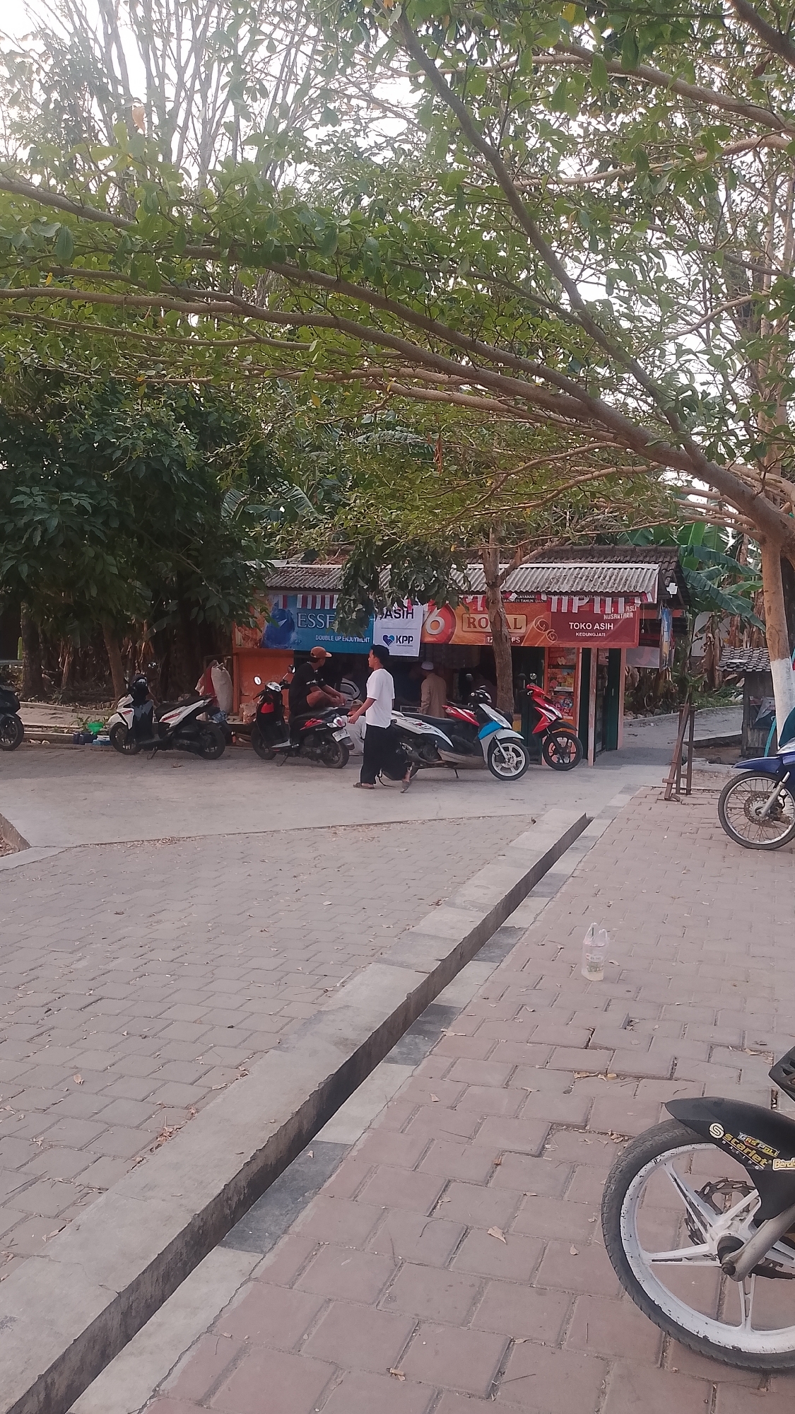 The image depicts a street scene in Karanglangu, Indonesia, under a canopy of trees. In the foreground, a paved area with interlocking brick pavers is visible, featuring a concrete drainage channel running along its length. A motorcycle is parked on the right side, showing its front wheel and part of the frame. In the mid-ground, several motorcycles are parked in front of a row of small shops with corrugated metal roofs. One shop displays the name "TOKO ASIH" and "KEDUNGJATI" in Indonesian. People are present near the shops; one man in a white shirt and black pants walks towards the right, while others are seated or standing in front of the establishments. The background is filled with trees and foliage, suggesting a semi-rural or suburban setting within the town. The lighting indicates it is daytime, with the sun casting shadows.