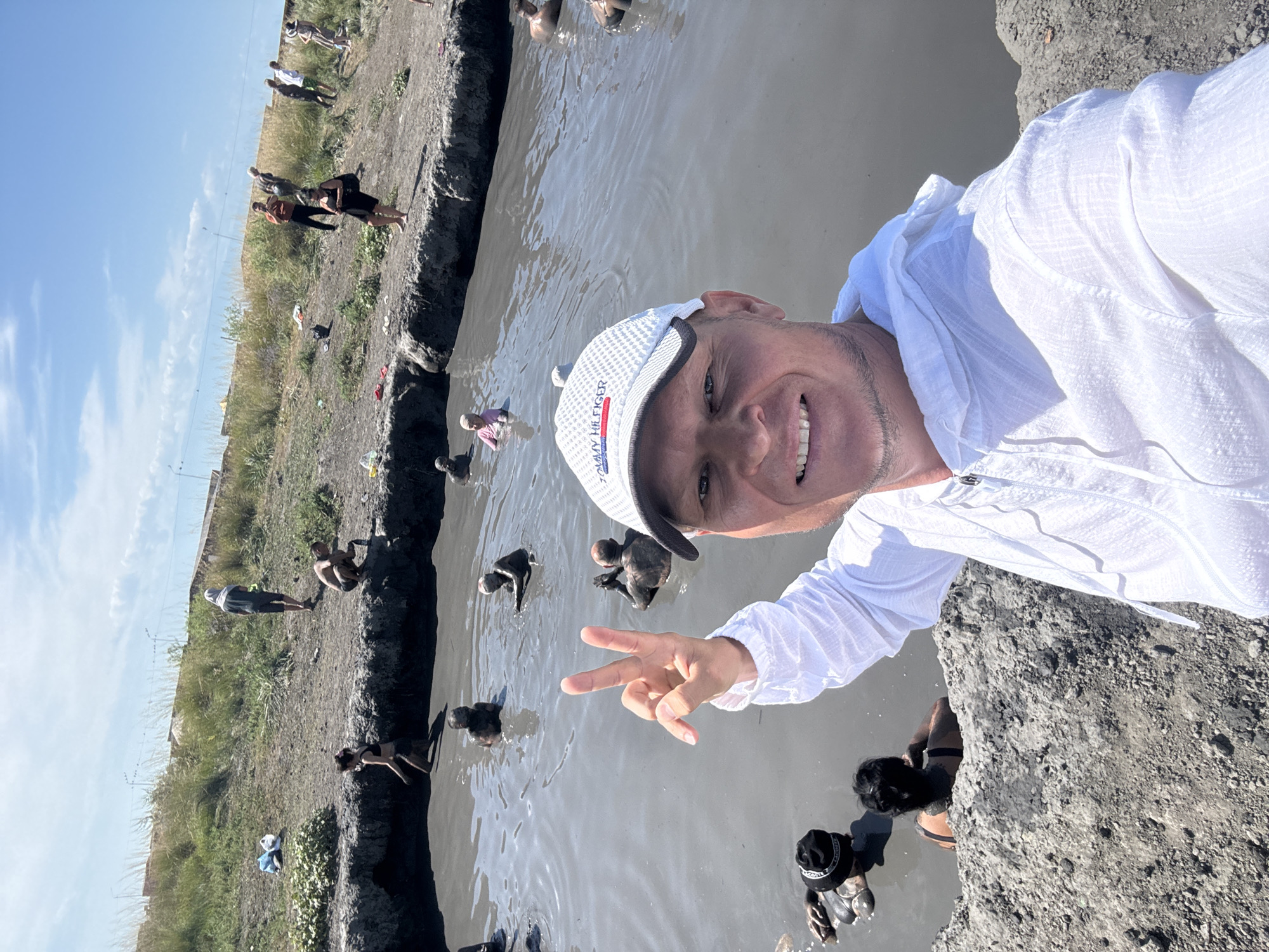 The image depicts a group of people bathing in a mud pool. In the foreground, a man wearing a white hoodie and a white baseball cap with "Tommy Hilfiger" written on it is taking a selfie. He is smiling and making a peace sign with his right hand. The mud pool is murky and grayish-brown, with several individuals immersed in it. Some are partially submerged, while others are mostly covered in mud. The surrounding environment consists of a barren, muddy embankment with dry grass and some scattered vegetation. In the background, there are more people standing on the embankment, observing the mud pool or preparing to enter it. The sky is clear and blue with some scattered clouds. The location is identified as a mud bath in the city of Zharbulak, Kazakhstan.