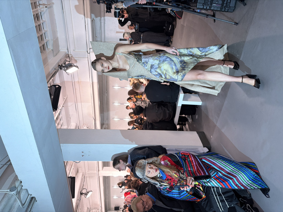 A fashion show scene captures a model in a patterned dress with a structured, asymmetrical overlay, confidently walking in black heels. Seated in rows, the audience intently watches, including a woman wrapped in a colorful, striped blanket and wearing a fur hat. Camera operators at the top document the event. The setting appears to be a bright, industrial space with exposed ceilings and structural supports.