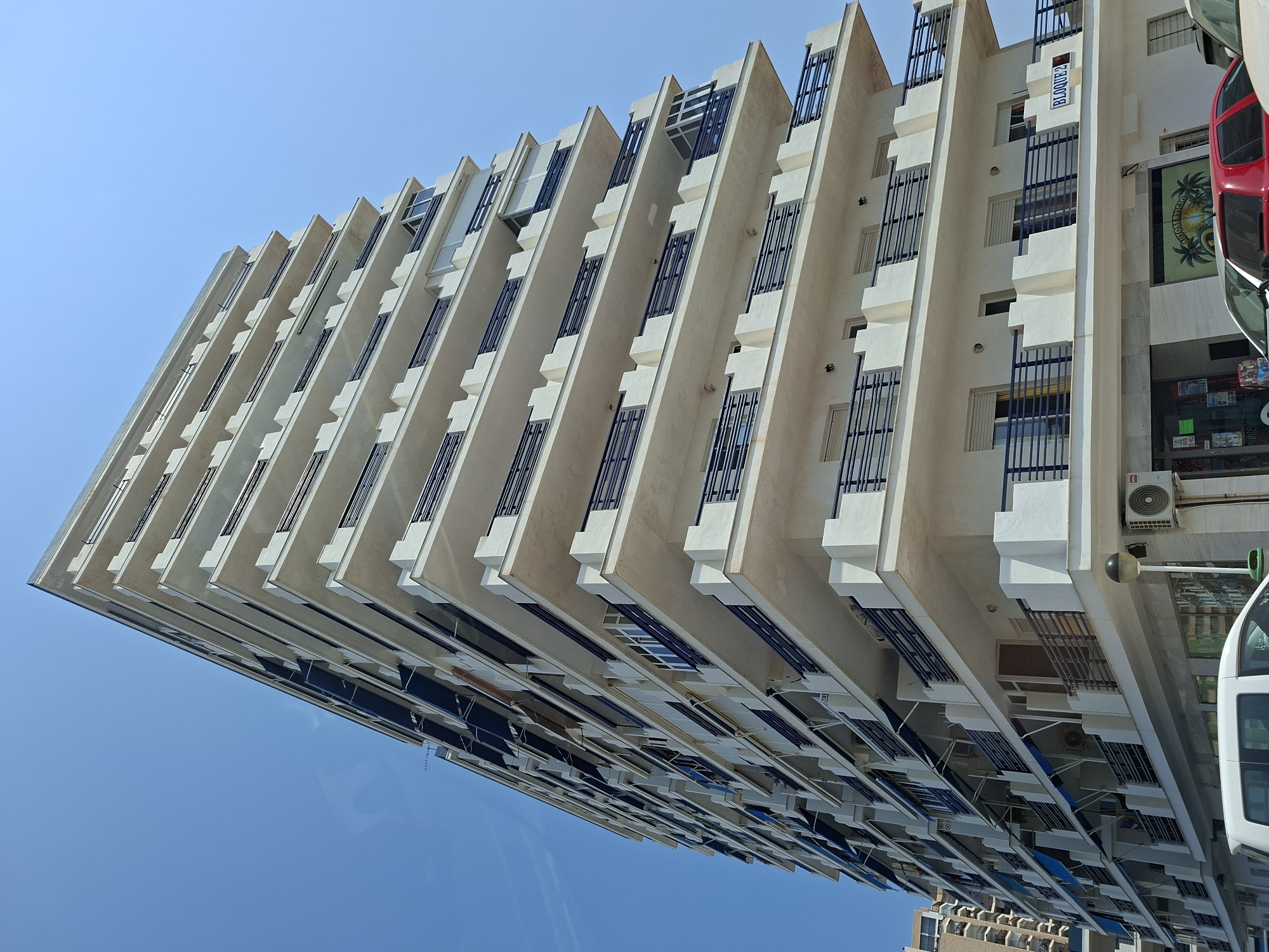 This is a low-angle, eye-level, medium shot of a modern, multi-story apartment building in La Manga Del Mar Menor, Spain, on a clear, bright, sunny day with a vivid blue sky. The building is tall and rectangular, with a white facade and numerous balconies. Each balcony is lined with dark blue railings, and many have retractable blue awnings extended. The architecture is characterized by repeated geometric shapes, with projecting elements on the balconies creating a layered effect. The lower floors appear to house commercial spaces, with storefront windows visible. One sign on the building reads "BLOQUE 2". There are air conditioning units mounted on the exterior walls. In the foreground, the top portion of a white van is visible on the left, and a red car is parked on the right. The overall impression is one of a densely populated coastal urban environment.