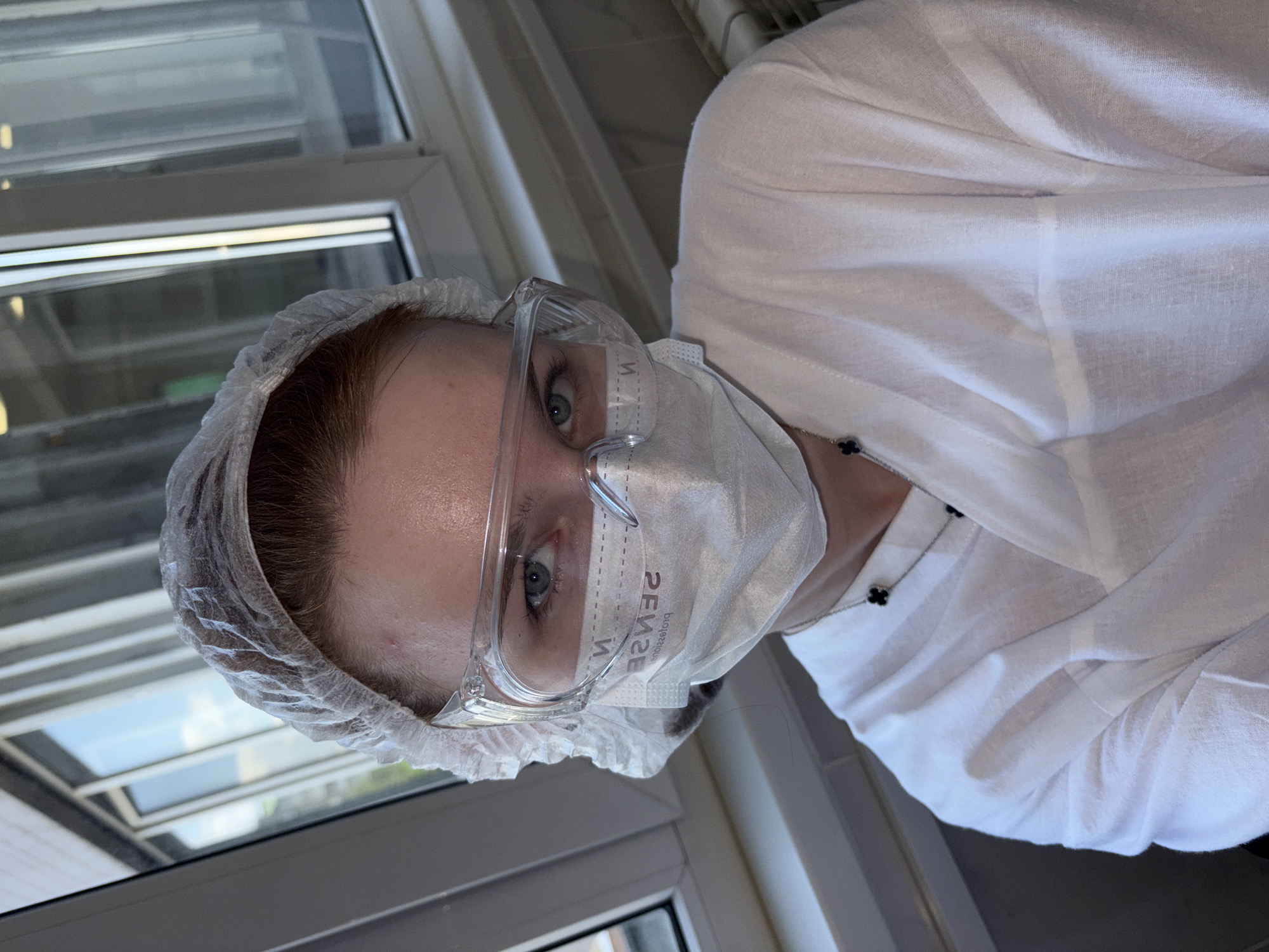 A portrait-style image, taken indoors, features an individual wearing protective gear. The person, who appears to be female, looks directly at the viewer with light blue eyes visible above a white disposable face mask. They are also equipped with clear safety goggles and a white disposable hairnet covering their head. A white long-sleeved garment, likely a professional shirt or lab coat, is worn, and a silver chain necklace with a black four-leaf clover pendant is visible at the collar.
The background shows large windows with white frames, through which natural daylight streams in, revealing a clear sky and reflections of what might be an exterior building or landscape. The overall setting suggests a professional or clinical environment, such as a medical office, laboratory, or specialized service facility, indicated by the hygiene and safety attire.
The face mask prominently displays the word "SENSE" (reversed) and partially visible smaller text that reads "professio" (also reversed). The scene appears to be lit by daylight, indicating it's daytime with clear weather. The individual's direct gaze conveys a neutral or observant expression. This scene is set in Saint Petersburg, Russia.