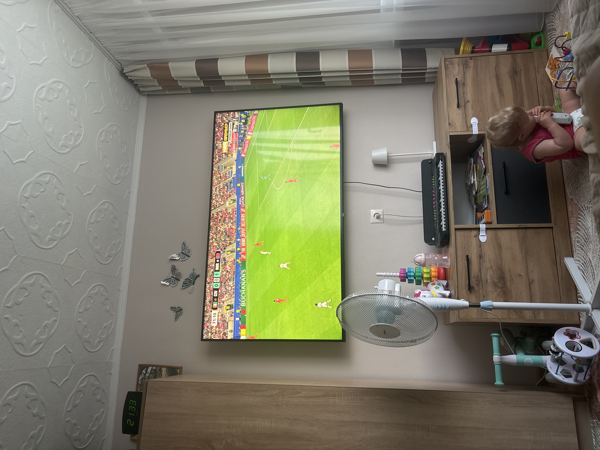 This media file captures an indoor scene, likely a living room in Krāslava, Latvia. The primary focus is a large television screen displaying a soccer match, indicated by the score "ClV 0 1 NOR" and the text "МАТЧ ПРЕМЬЕР" and "ПРЯМОЙ ЭФИР" which translates to "MATCH PREMIER" and "LIVE BROADCAST." The time displayed on a digital clock on a wooden cabinet reads "21:33," suggesting it is evening.
To the right of the television, a young child with blonde hair, wearing a pink shirt and a diaper, is seated on a beige surface, possibly a bed or sofa. The child appears engaged with a white object, possibly a toy. Next to the child is a wooden cabinet containing various items, including children's toys and bottles. A white oscillating fan is positioned in front of the cabinet, and other toys are visible on the cabinet and nearby shelves. Decorative butterfly decals adorn the wall to the left of the television. The room is illuminated by artificial light, and the presence of a digital clock and a television suggests a domestic setting.