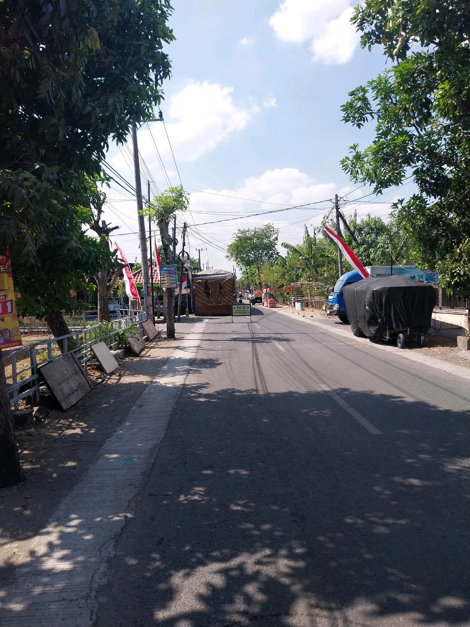 The image shows a street scene in Campur, Indonesia, on a sunny day with a clear blue sky and scattered clouds. The road is asphalt with a white line marking the center. On the right side of the road, there is a blue truck covered with a black tarp, and next to it, a cart with blue wheels. On the left side of the road, there is a concrete sidewalk and a metal fence. Several Indonesian flags are displayed on poles along the side of the road, indicating a festive or commemorative occasion. Trees with green foliage are visible on both sides of the road, casting shadows on the pavement. In the background, there are buildings and more vegetation. There are no people clearly visible in the frame.