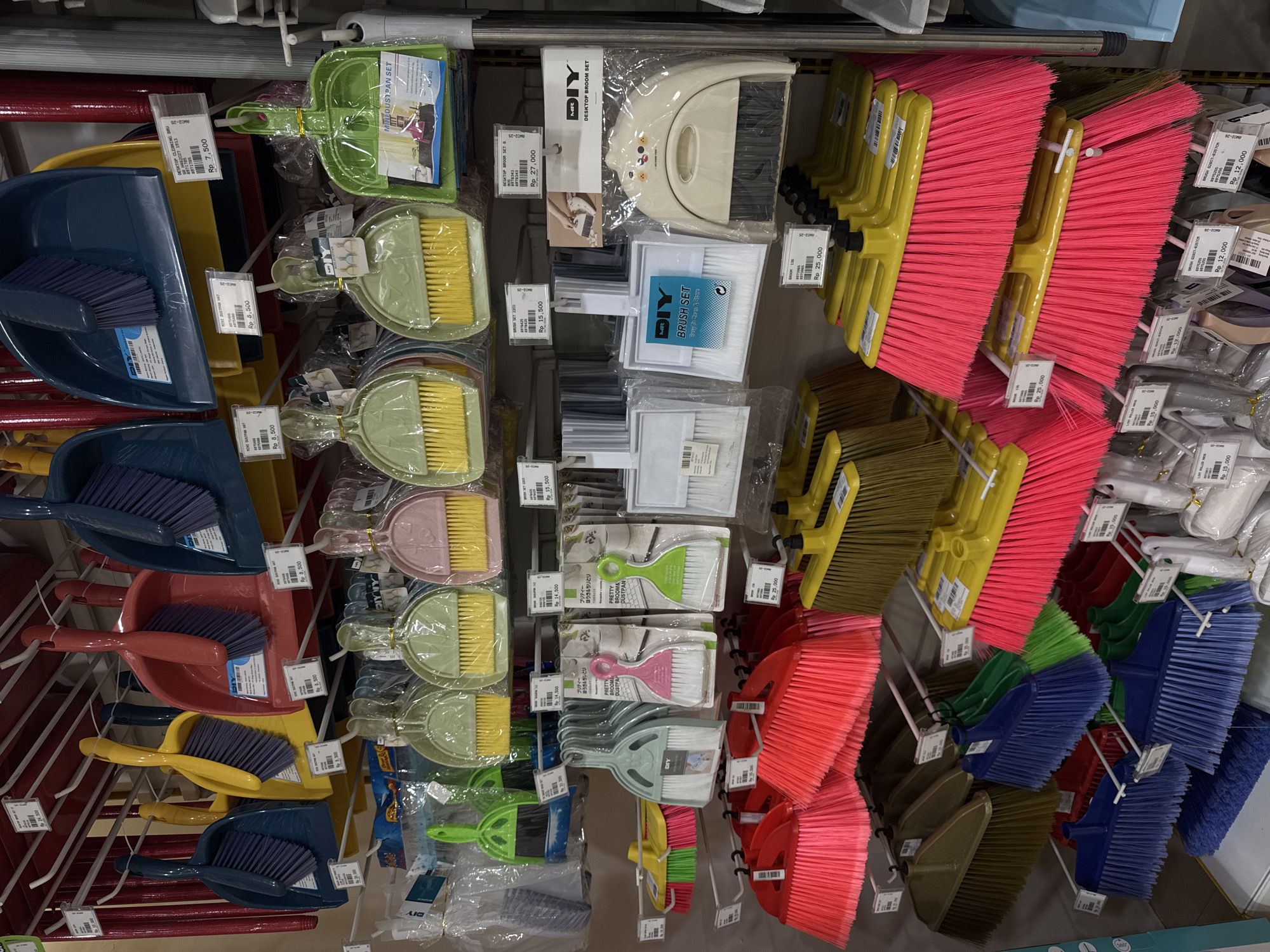 The image depicts a display of cleaning supplies in a retail setting. The items consist of a variety of brooms, dustpans, and desktop cleaning sets, arranged on shelves. Various price tags are affixed to the products. The price tags display Indonesian Rupiah amounts. The OCR text indicates a location of Kuta Selatan, Indonesia.