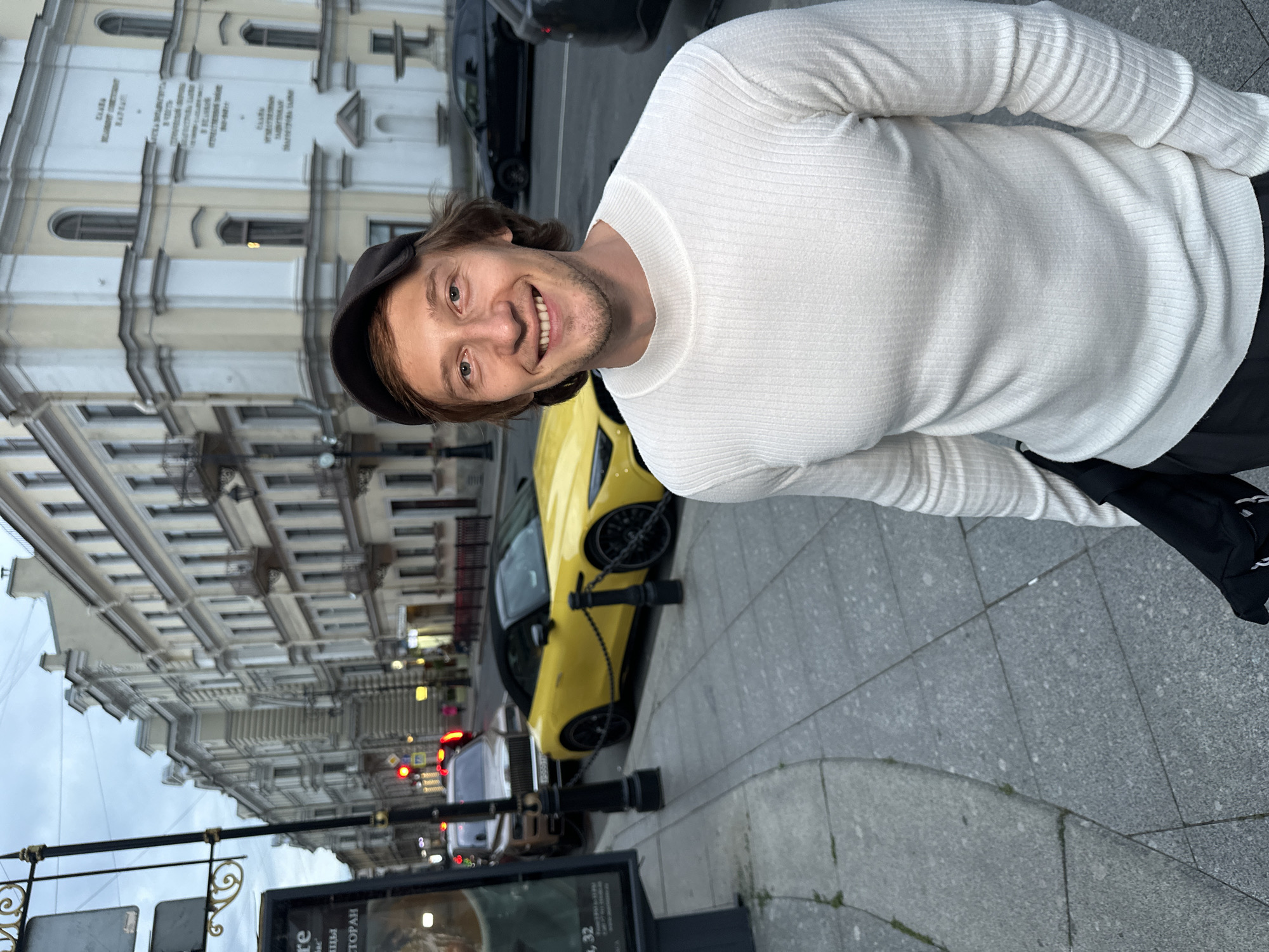 A man with light brown hair and a beard, wearing a white ribbed sweater and a dark baseball cap, smiles at the camera. He stands on a grey sidewalk in front of a yellow convertible sports car. In the background, a row of historic buildings lines a street in Saint Petersburg, Russia. A billboard is visible on the left side of the image. The sky is overcast.
