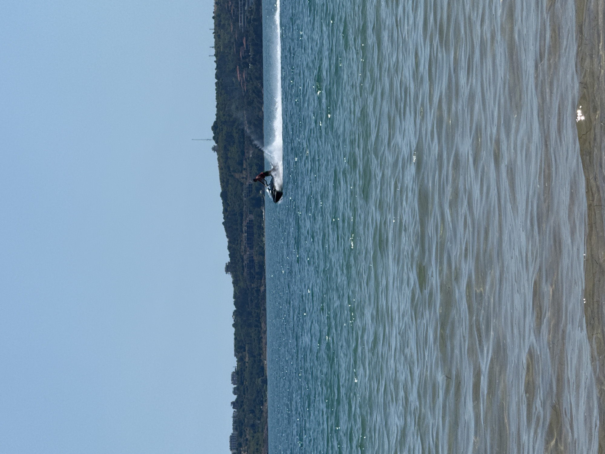 The image depicts a bright, sunny day at the coast of Sozopol, Bulgaria. The sea, with its deep blue color and gentle waves, occupies the majority of the frame, with the shore visible in the foreground. In the mid-ground, a person is riding a jet ski, leaving a white wake as they speed across the water. The background features a verdant, hilly landscape dotted with buildings, indicating a developed coastal area. The sky above is a clear, light blue.
