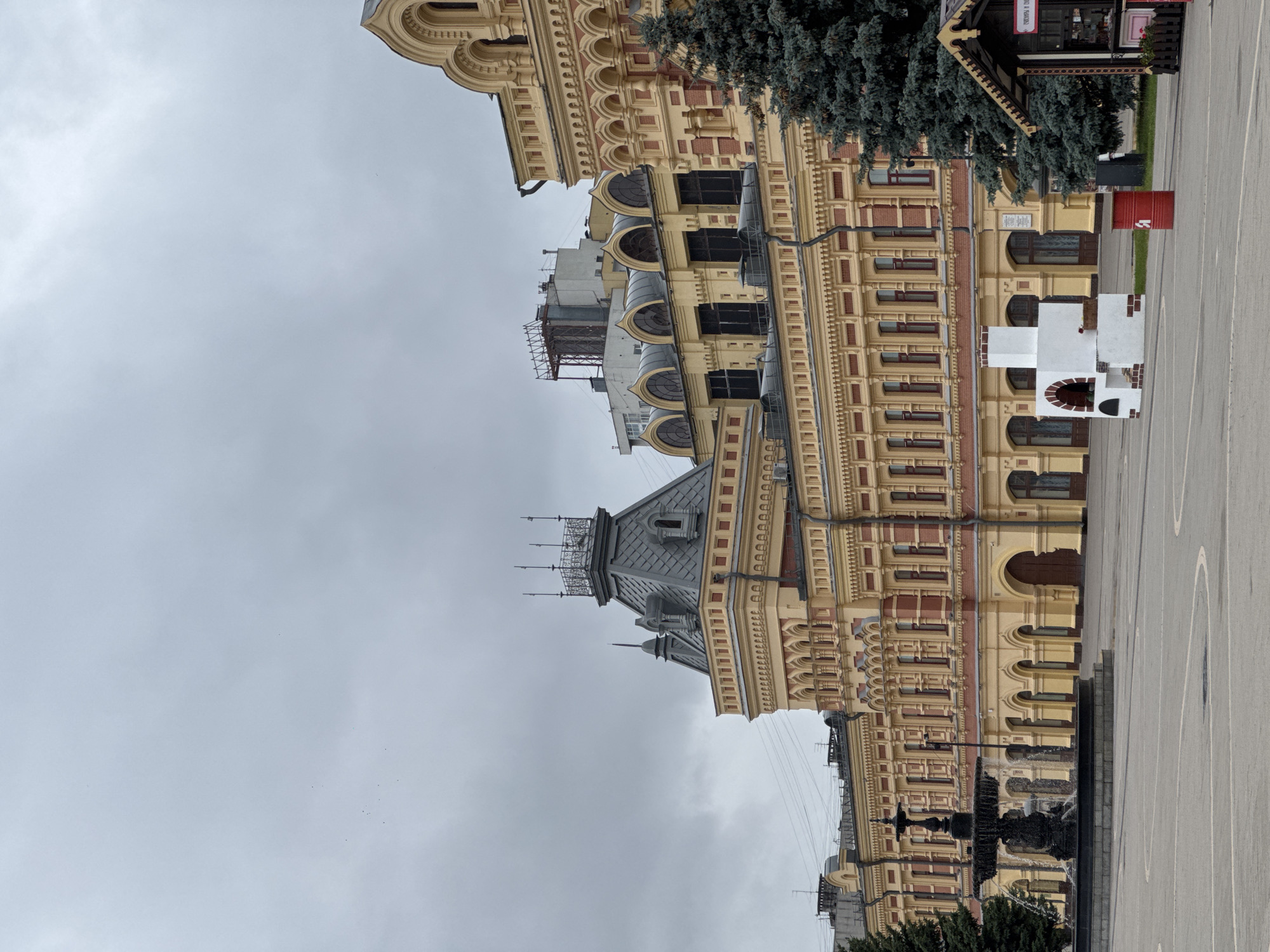 The image captures an outdoor scene in Nizhny Novgorod, Russia, likely on an overcast day. The main subject is a large, ornate yellow and red building with a distinctive architectural style featuring numerous arched windows and decorative elements. In the foreground, to the left, is a dark, multi-tiered fountain with water flowing. To the right of the fountain and in front of the building, there is a white, brick-like structure resembling a decorative oven or hearth, with a basket of red flowers placed inside. A red cylindrical trash bin with Cyrillic lettering is positioned near this structure. Adjacent to the oven, a small wooden kiosk with a peaked roof and decorative trim is visible, displaying items for sale and a sign with Cyrillic text. Several evergreen trees, appearing to be fir or spruce, are present in the mid-ground, partially obscuring the building. The ground is paved with light-colored cobblestones or tiles, with visible white lines delineating a plaza area. The sky is overcast and a pale gray. In the upper left portion of the sky, a few small dark specks that could be birds are faintly visible.