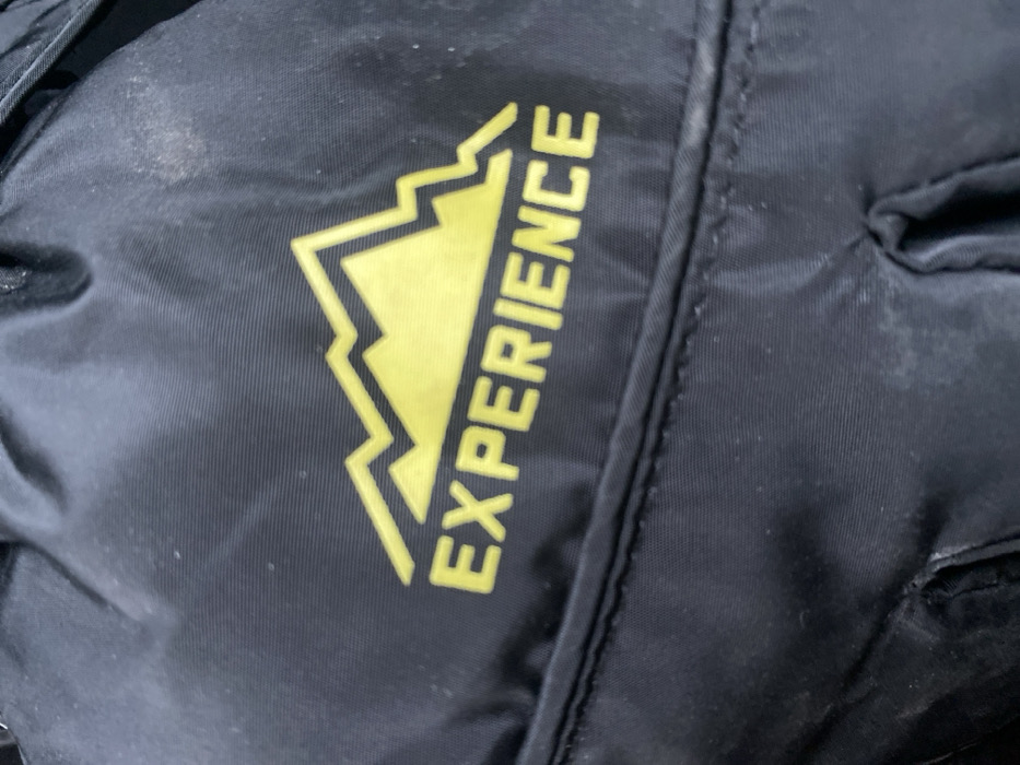 A close-up image displays a section of black synthetic fabric, consistent with material found on a backpack or outdoor gear, located in Pavlovo Podvor'ye, Russia. A yellow graphic is prominently featured, depicting a stylized mountain range above the capitalized word "EXPERIENCE." The fabric shows minor surface accumulation of light-colored dust particles, primarily noticeable in the upper right quadrant and along the bottom edge. A horizontal stitched seam is visible beneath the "EXPERIENCE" text, and the material exhibits slight creasing in various areas. No individuals or specific actions are depicted.