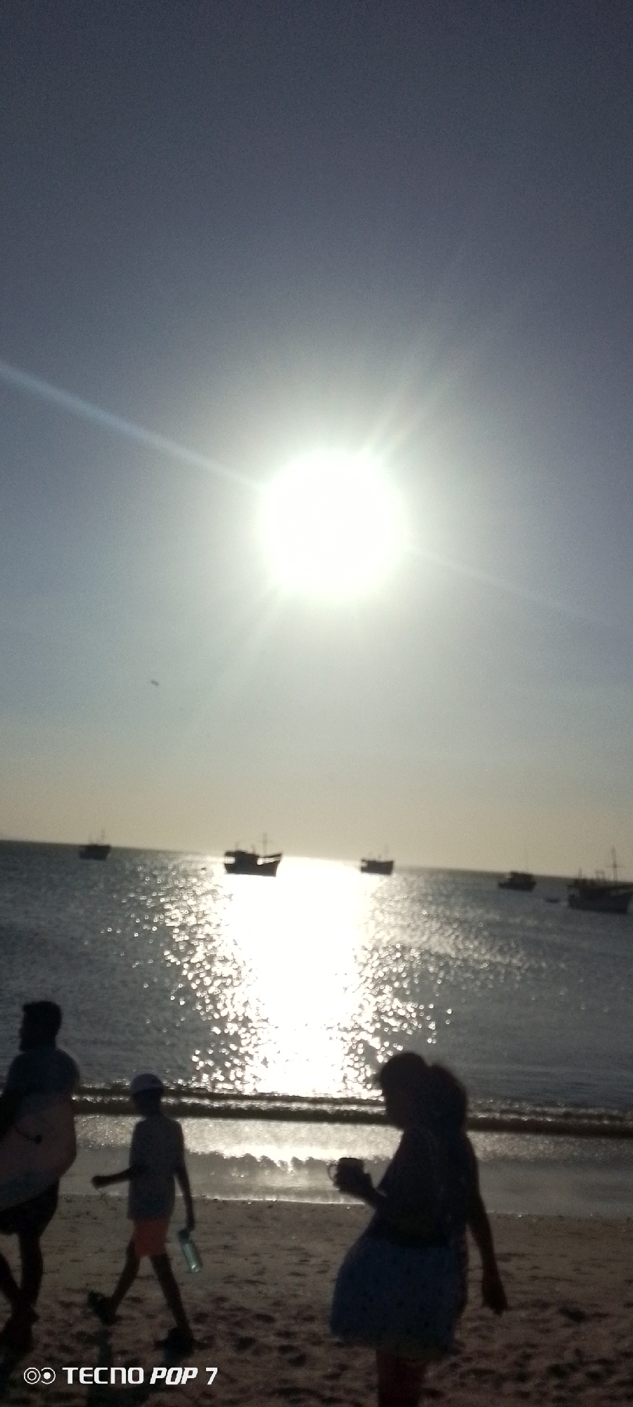 The photograph captures a bright scene at a beach, possibly in Altagracia, Venezuela, where the sun shines intensely over the ocean. Several boats are visible on the water in the distance. In the foreground, indistinct figures of people can be seen walking on the sand near the shoreline. The intense sunlight creates a strong glare and silhouettes, making it difficult to discern details about the individuals or the environment.