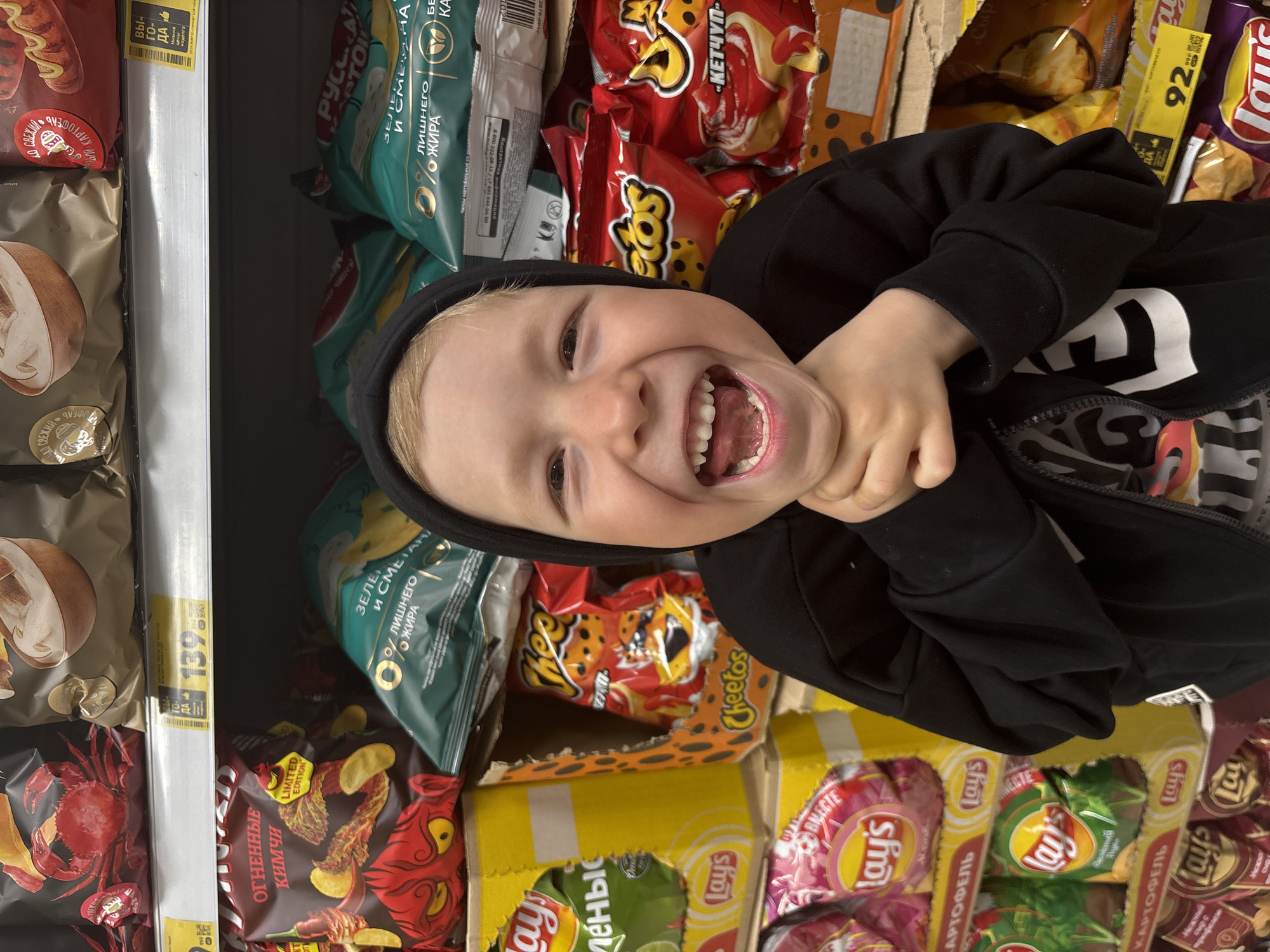 A young boy with blond hair, wearing a black hooded sweatshirt over a patterned shirt, is smiling widely at the camera. He is surrounded by stacks of chips in a retail setting. Visible chip brands and flavors include Lay's (various flavors in Russian), Cheetos "Ketchup," and "Russian Potato" chips in different flavors. A price tag of 139 is visible near chips with mushroom and crab imagery. The chips are on shelves in a retail store in the city of Chaikovsky, Russia.