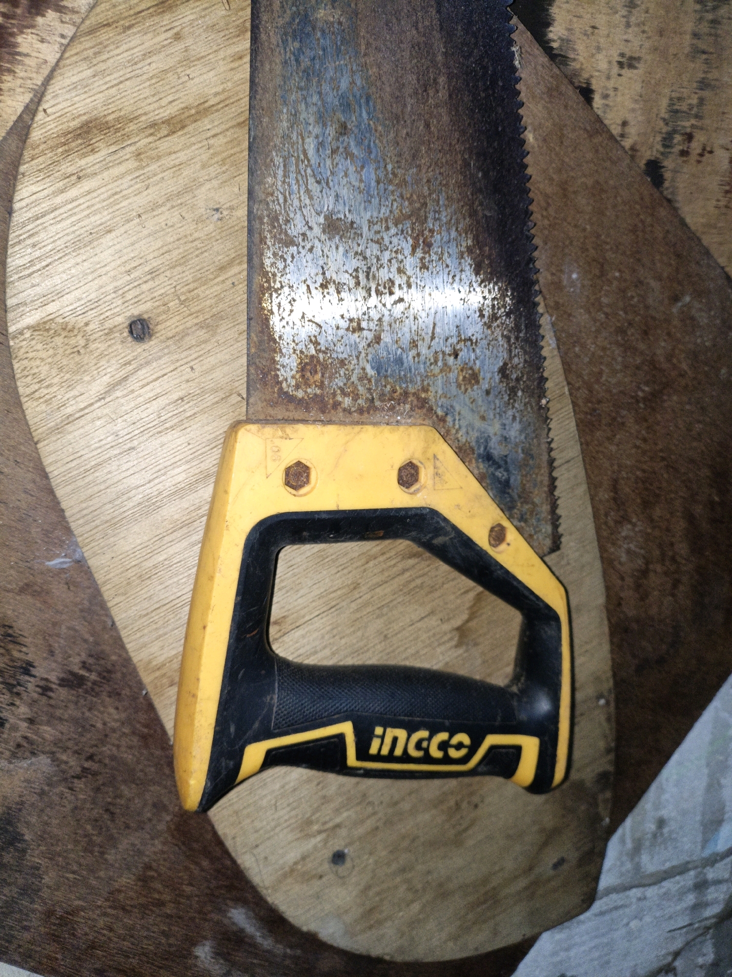 The image shows a close-up of a rusty hand saw with a yellow and black handle lying on a wooden surface. The saw blade is heavily corroded, with visible patches of rust and discoloration. The handle is made of plastic and features the "ingco" logo. The wooden surface appears to be part of a larger object, possibly a workbench or a sheet of plywood, with visible wood grain and some imperfections. The lighting suggests an indoor environment, possibly a workshop or storage area. There is no information about the specific location in Cikal, Indonesia, within the provided image.