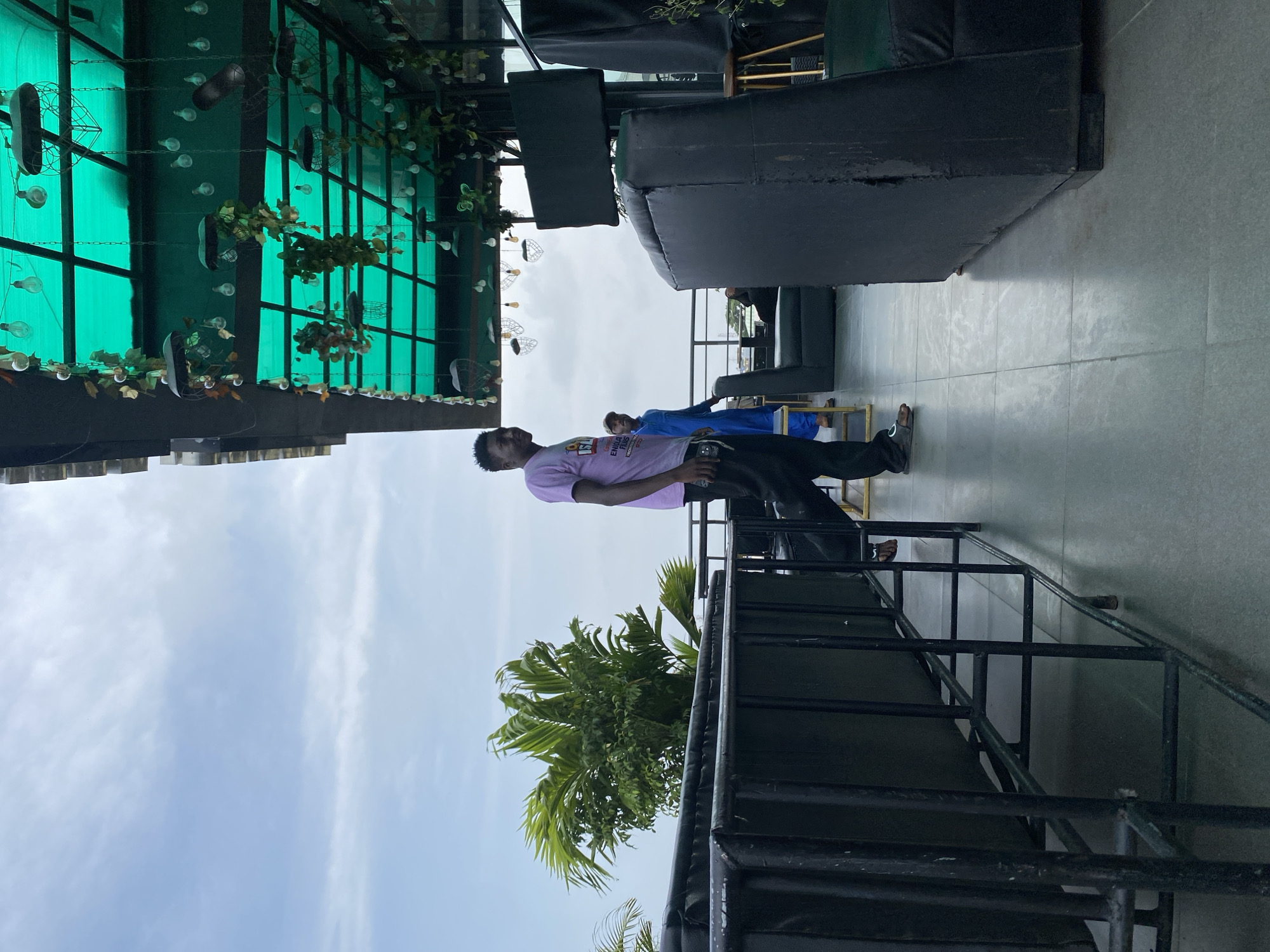 An outdoor rooftop seating area at a restaurant or bar is shown. The space features black metal railings, dark upholstered seating, and a green translucent roof with hanging decorative lights and artificial greenery. A young man wearing a pink t-shirt and black pants stands facing left, looking towards the side. Another person in a blue outfit is visible further back in the seating area. Palm trees are present in the background, suggesting a tropical or warm climate. The sky is overcast.