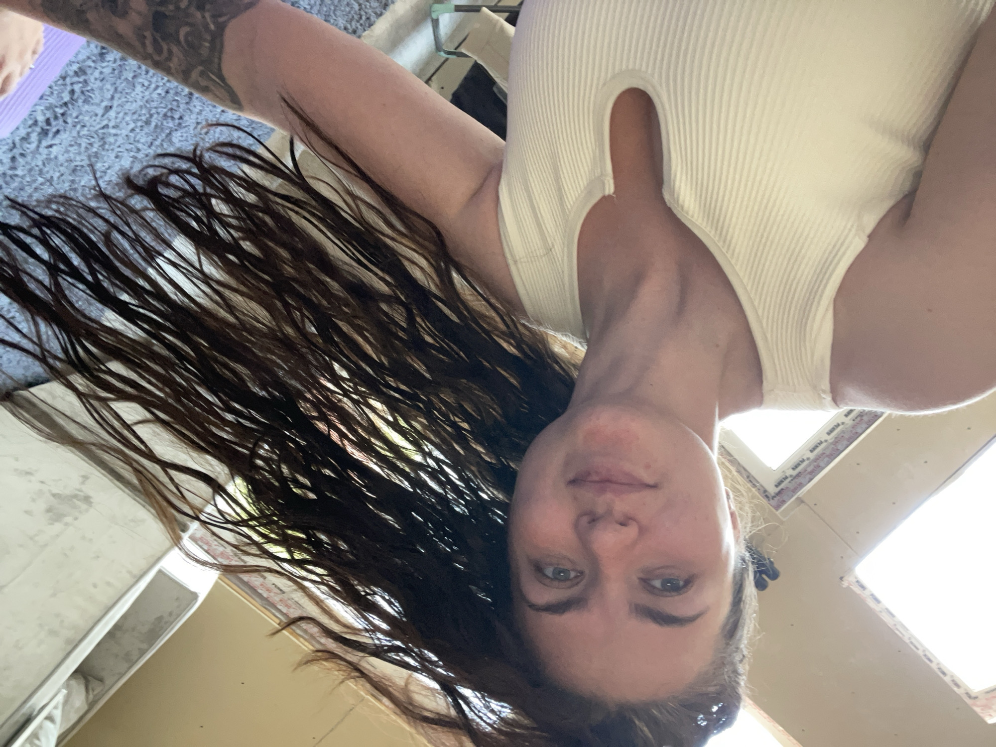 A woman with long, dark, wet hair is photographed from below, looking directly into the camera. She is wearing a white, ribbed tank top with a keyhole neckline. Her arms are visible, with a tattoo on one arm. The background includes a gray textured surface, possibly a rug or carpet, a glimpse of what appears to be part of a yoga mat in purple, and glimpses of interior structures with windows and light coming through them. The location within the city of Мосметаллоконструкция Онт, Russia, cannot be determined from the image.