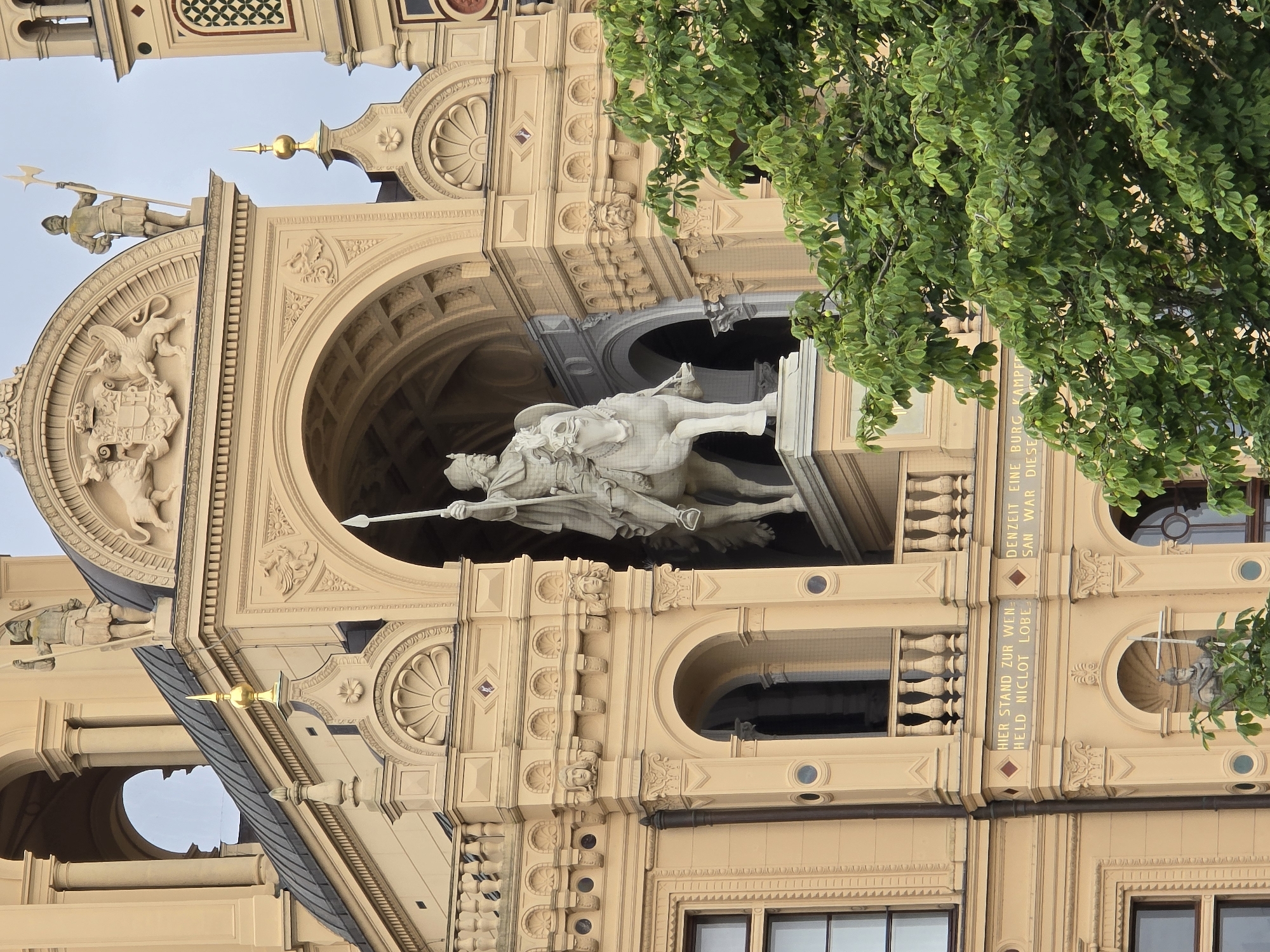 The image shows a close-up, eye-level view of the exterior of Schwerin Castle in Schwerin, Germany. The castle is an elaborate structure with a light yellow facade adorned with classical architectural elements. In the center of the frame, an equestrian statue of a man in armor, holding a spear, is prominently displayed within an arched alcove. The statue is made of a light-colored material, likely stone or marble. Above the statue, decorative friezes featuring coats of arms and mythological creatures are visible. Statues of armored figures holding weapons stand on top of the structure.
The castle's architecture includes multiple arches, columns, balconies with ornate railings, and windows. Below the equestrian statue, an inscription in German is visible on a band across the facade. A lush green tree with abundant leaves partially obscures the lower right portion of the castle. The sky appears overcast. The image focuses on the detailed ornamentation and the imposing presence of the castle's architecture and statuary.