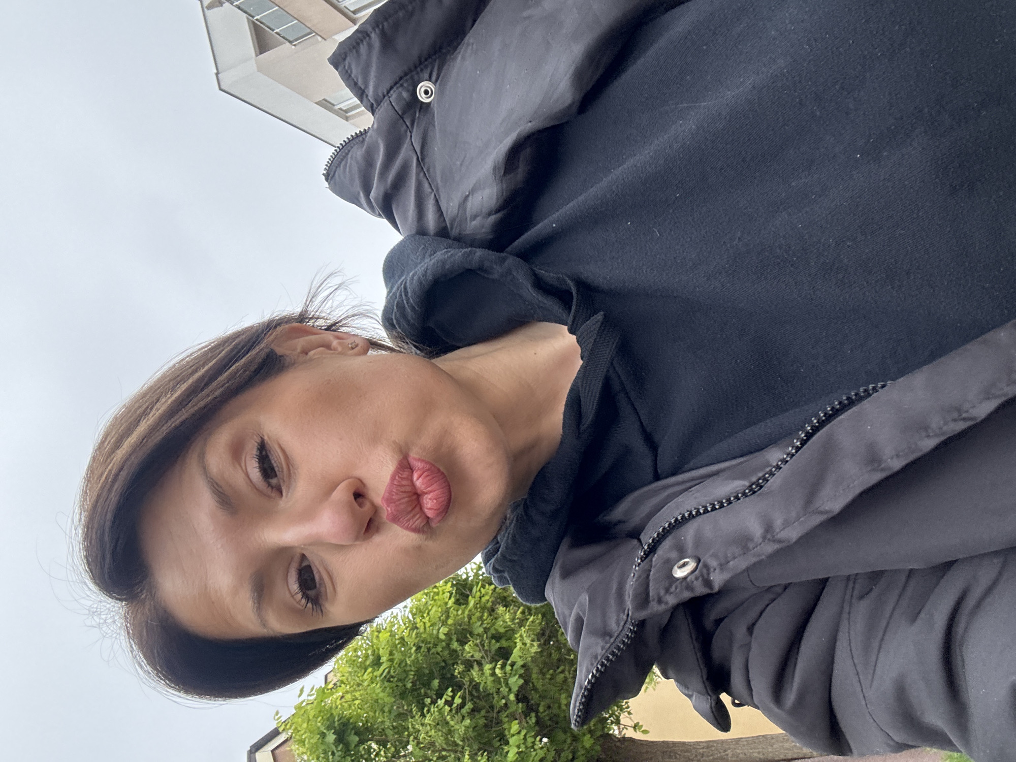 A woman with medium-length brown hair is taking a selfie. She has brown eyes, fair skin, and is pouting her lips. She is wearing a black hoodie and a black jacket. The background includes a building with windows and a green tree. The sky is overcast. The location may be in Sofia, Bulgaria, but it is not definitively discernible from the image.