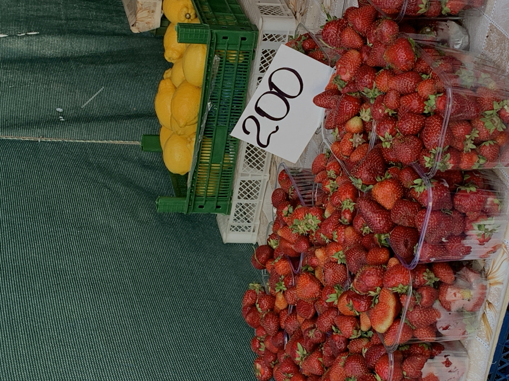 The image depicts a market stall, likely in Kyiv, Ukraine, displaying produce for sale. A green fabric-covered surface occupies the left side. To the right of this, are stacked green and white plastic crates. The green crate is filled with yellow lemons. Adjacent to these crates is a white card with the handwritten number "200" in large black numerals. Next to the sign are several transparent plastic containers stacked with red strawberries. The containers of strawberries dominate the right side of the image, overflowing with fruit. The image is captured from a high-angle perspective.