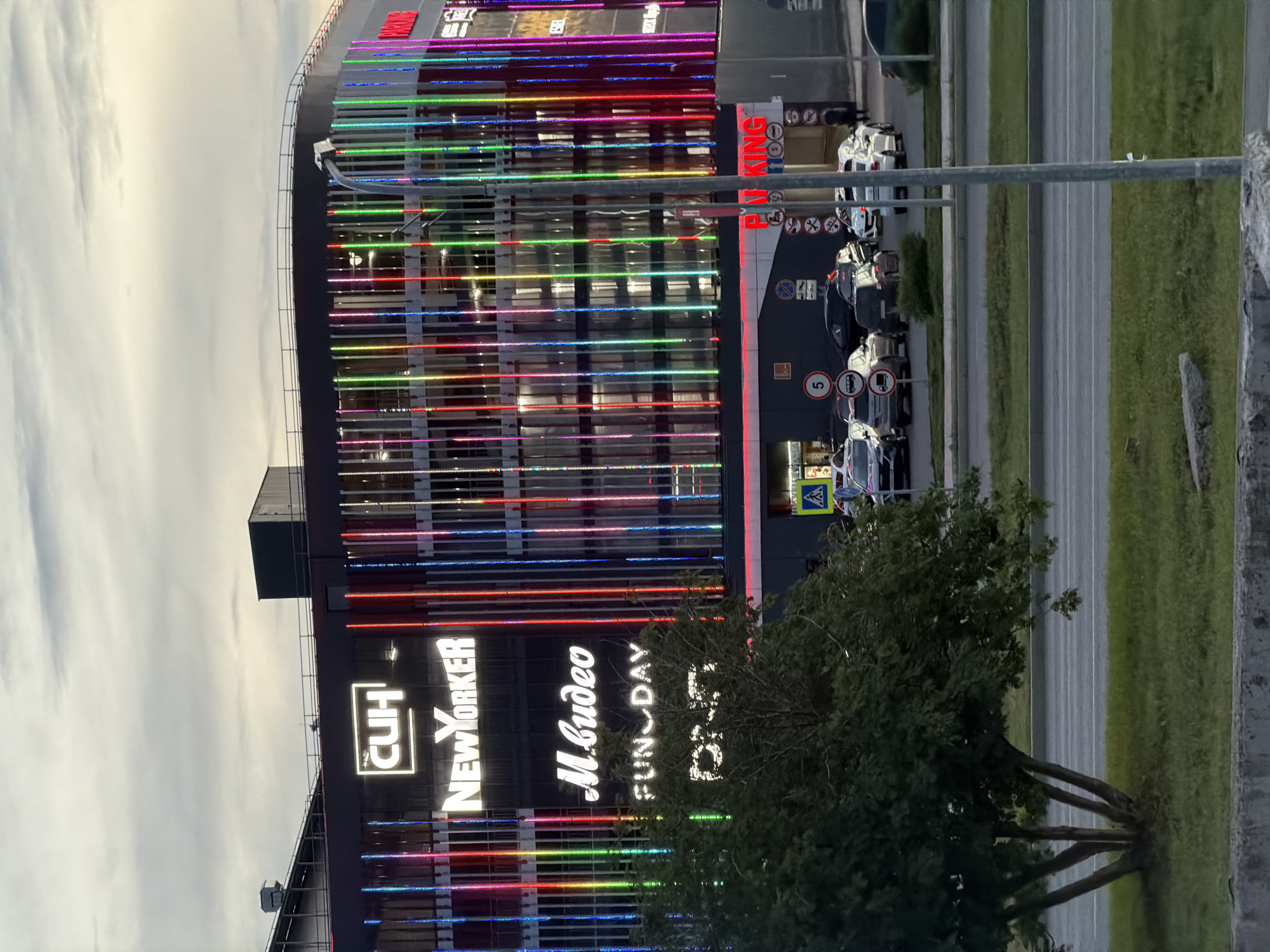 This media file captures a nighttime or dusk scene of a modern building with a brightly lit facade adorned with colorful LED lights. The building appears to be a commercial establishment, possibly a shopping center or entertainment venue, as indicated by various signs. Text visible on the building includes "PARKING," "CUH," "NEW YORKER," "M.видео," "FUN DAY," and "DIET." Several cars are parked in front of the building, suggesting it is open to the public. A tree with dense foliage stands in the foreground, partially obscuring the lower portion of the building. The sky is overcast, and the overall lighting suggests a twilight or evening setting. The scene appears to be located in Nizhnekamsk, Russia, given the provided location context. No people are clearly visible in the frame, but the presence of cars and the illuminated building imply ongoing activity. The vibrant, multi-colored lights on the building create a festive and dynamic visual effect against the darkening sky. The overall impression is one of a lively urban environment, likely a popular destination for shopping and entertainment.