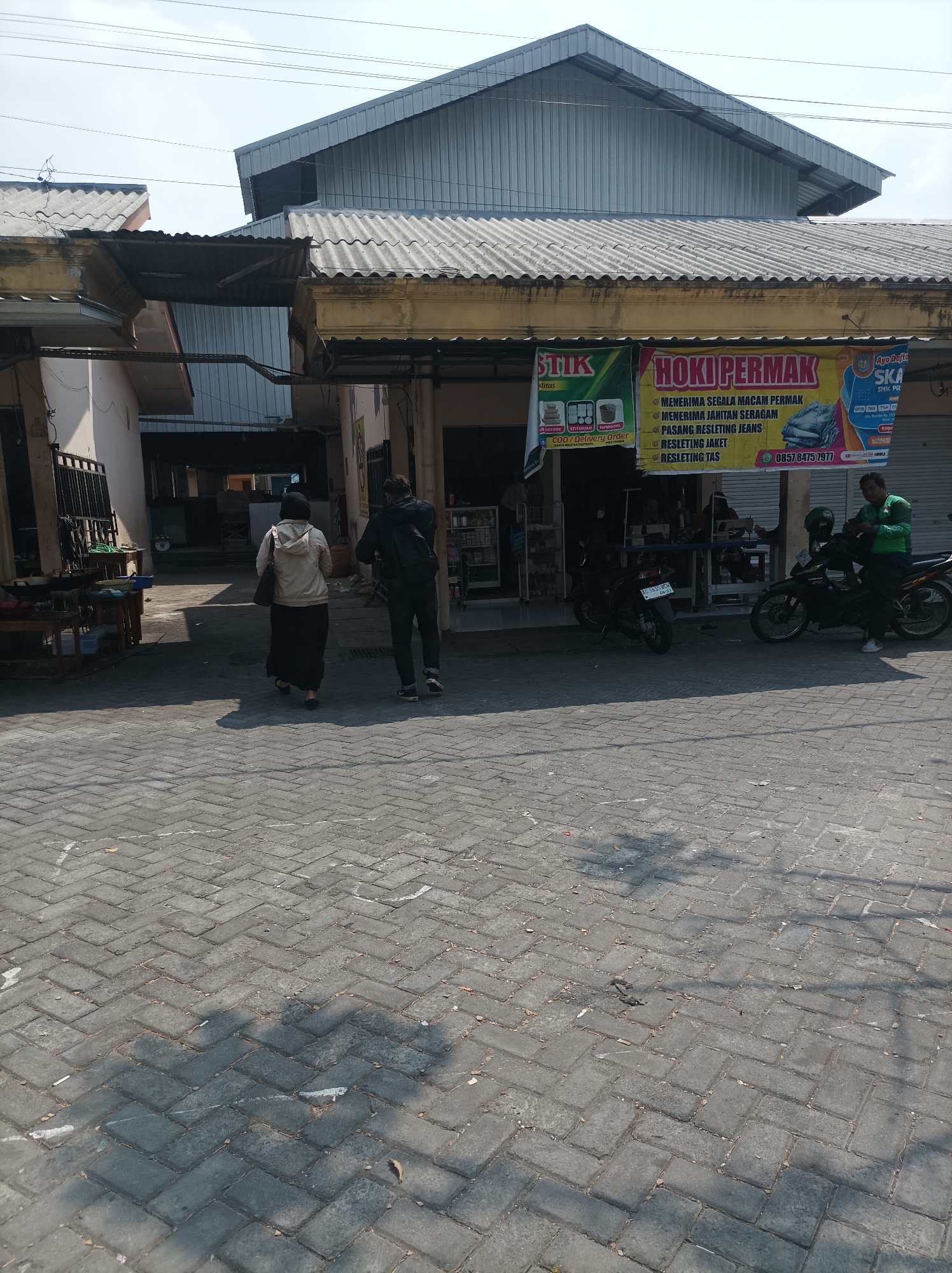 The image captures a street scene in Nganjuk, Indonesia, likely in front of small businesses. In the foreground, a paved area with herringbone brickwork is visible, marked with white paint lines and scattered debris. Two individuals are walking away from the camera on this paved area. To the right, a parked motorcycle with a rider wearing a green shirt is present.
Behind them, a row of buildings features corrugated metal roofs and yellowed facades, suggesting some age. Several shops are visible, with one displaying banners that advertise tailoring and alteration services, including the text "HOKI PERMAK" and phone numbers. A motorcycle is parked in front of one of these shops. Further back, the street continues, with more buildings and what appears to be an alleyway or a passage between structures. The overall atmosphere is that of a typical commercial area in a moderately busy Indonesian town. The time of day appears to be daytime, with clear skies and distinct shadows cast by the buildings and the people.