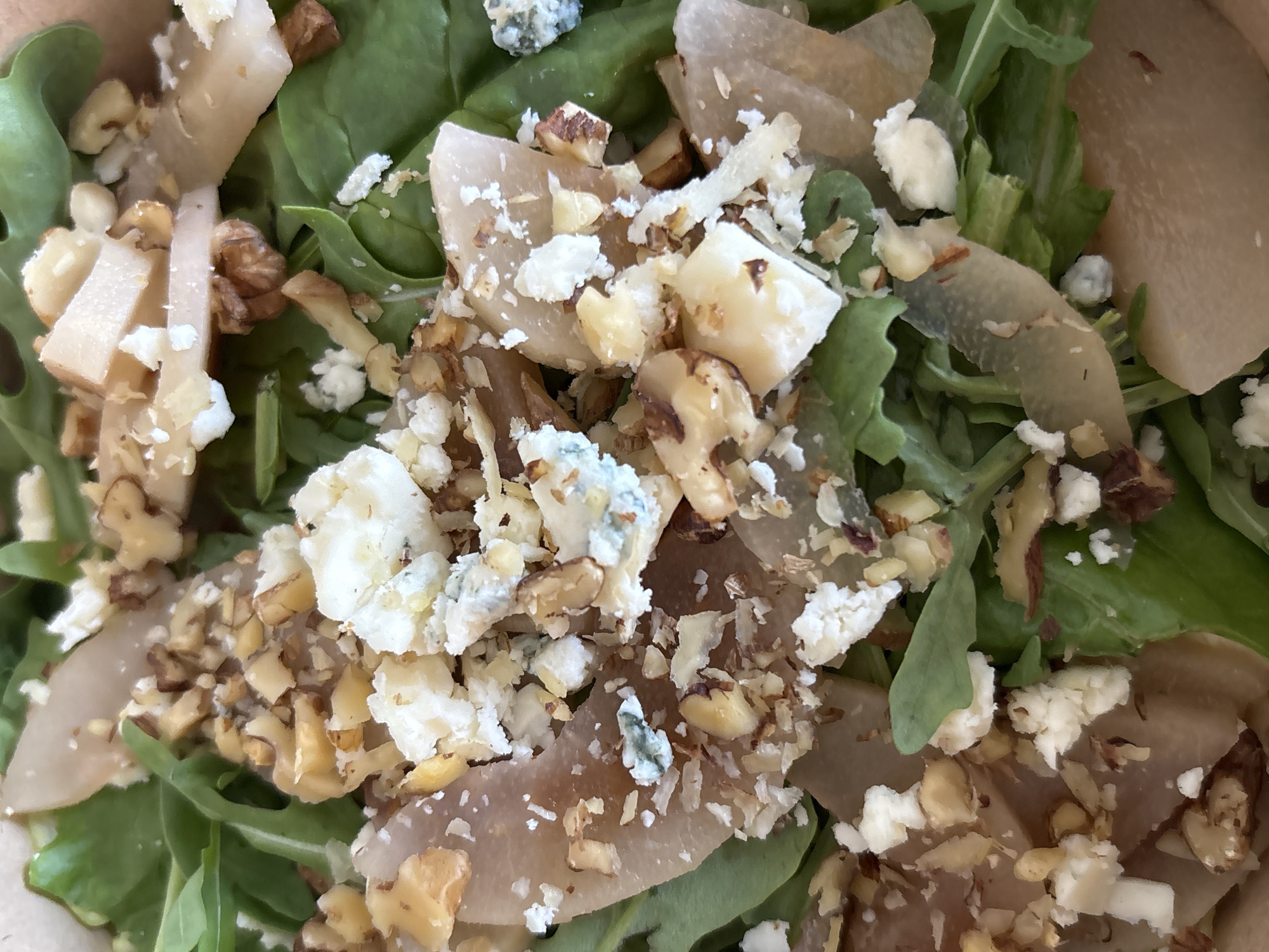This is a close-up, overhead shot of a salad. The salad appears to be a mix of leafy greens, thinly sliced pears, crumbled cheese (likely blue cheese), and chopped walnuts. The ingredients are artfully arranged in a shallow container, possibly a takeout bowl. The lighting is natural and bright, highlighting the textures and colors of the salad. There are no people or any identifiable background elements that would suggest a specific location within Shushary, Russia. The image focuses solely on the food itself, presenting it as a visually appealing and fresh dish.