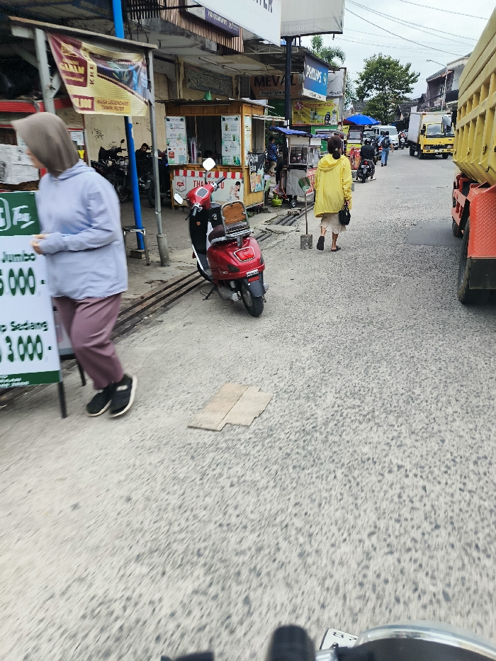 The image shows a street scene in Kecamatan Dayeuhkolot, Indonesia. A woman in a grey hoodie and pink pants is walking towards the camera while holding a sign with the words "Jumbo 5.000" and "Sedang 3.000". In the background, a woman in a yellow jacket walks away from the camera. A red scooter is parked on the right side of the street, and a large truck is parked on the far right side of the street. A sign with the word "Meval" is visible in the background. The street is paved with small gravel stones. There are buildings on the left side of the image and the sky is overcast. The street is relatively empty, with only a few people and vehicles present.