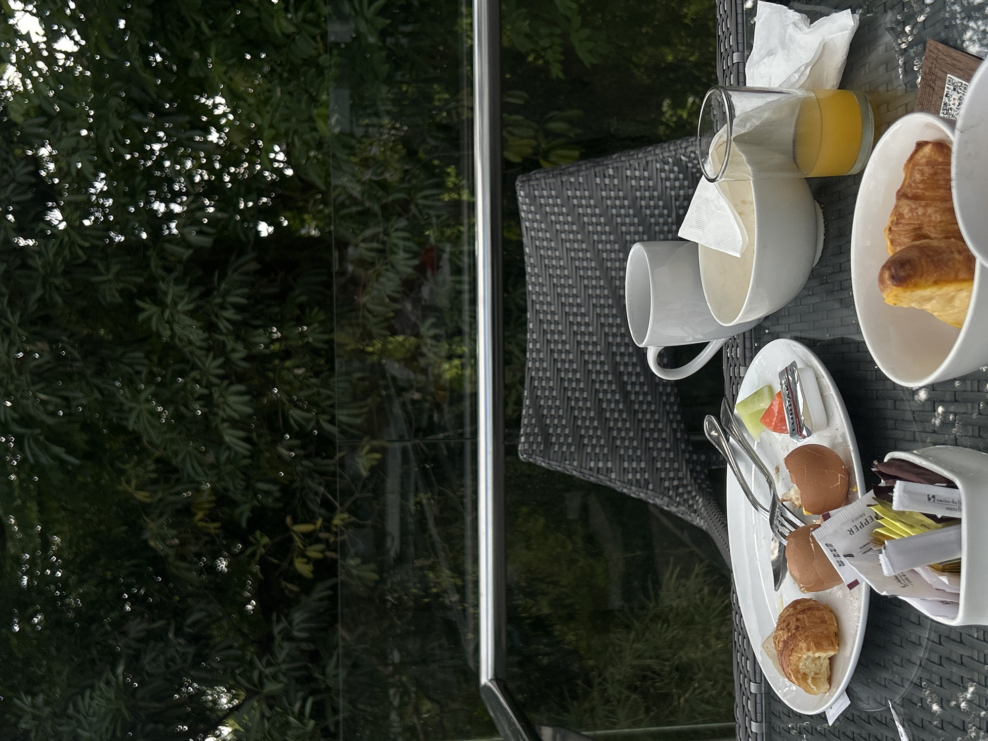 The image shows a table with breakfast items, set against a backdrop of lush green foliage and a glass railing. The table is laid out with a white plate containing a half-eaten croissant, two boiled eggs in their shells, a small packet of butter, and some cut fruit. A fork and knife rest on the plate. In front of the plate is a white bowl filled with sachets of sugar and artificial sweetener. To the right of the plate, a white mug sits next to a white bowl. Further to the right, a glass of orange juice is visible, alongside a crumpled paper napkin. In the foreground, another white bowl contains two more croissants. The overall setting suggests an outdoor dining area, likely a balcony or patio, with trees and vegetation forming a natural screen behind the railing. The lighting suggests it is daytime, possibly morning given the breakfast setting. The scene is devoid of any people. The location is identified as Kuta Selatan, Indonesia.