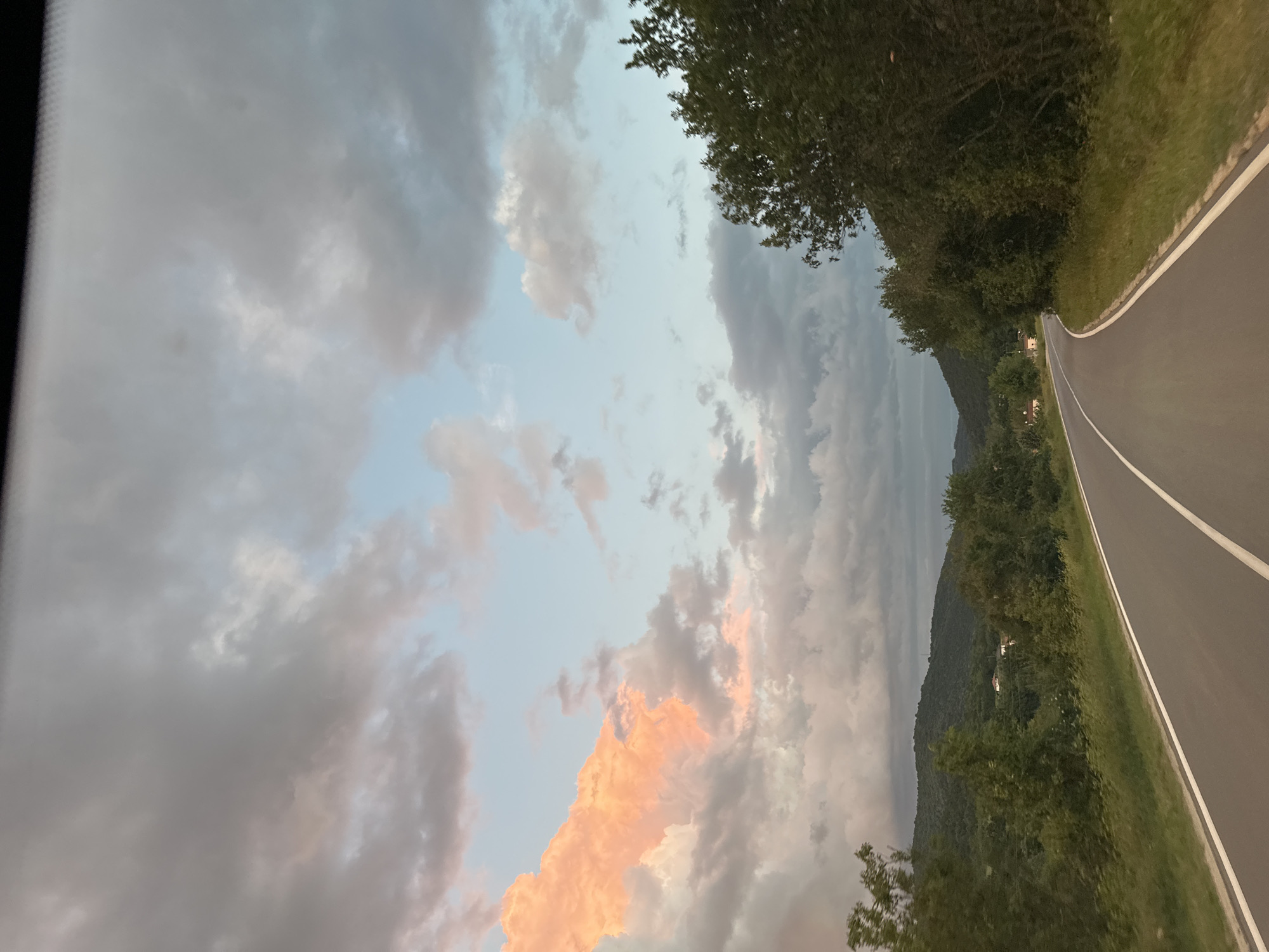 The image depicts a view from inside a vehicle, likely a car, driving on a road. The road is asphalt with a white line marking the edge. Trees line the right side of the road, obscuring a view of the landscape beyond, which appears to include a hill or mountain. The sky dominates the left side of the frame, showing a mixture of clouds, some lit with a warm orange glow from the setting sun. The overall scene has a slightly overcast or dusky quality.
The specific location, Pryaporets, Bulgaria, is not directly discernible from the visual information.