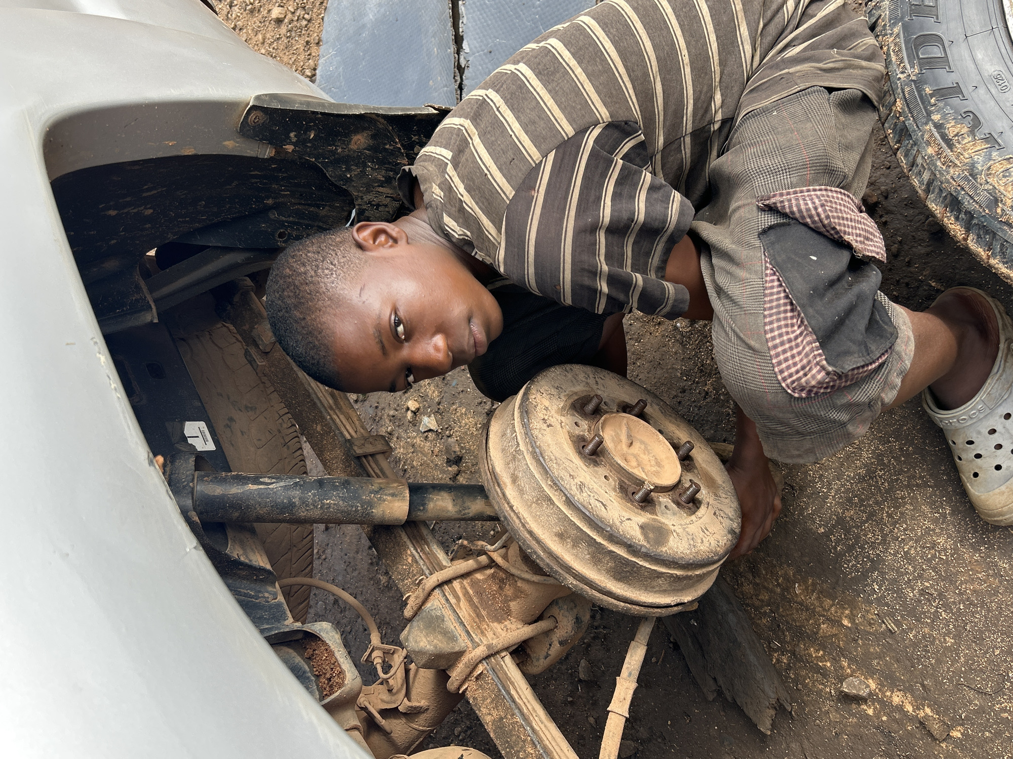 A young boy is shown kneeling next to the rear axle of a vehicle, seemingly working on the brake drum. He is wearing a striped t-shirt and checkered shorts, and appears to be in an outdoor setting that suggests a workshop or roadside repair area, possibly in Barikin Jirga Summa, Nigeria. The ground is covered with dirt and debris, indicative of a working environment. The vehicle itself appears to be older, with visible signs of wear and tear. The boy is looking directly at the camera with a serious expression.