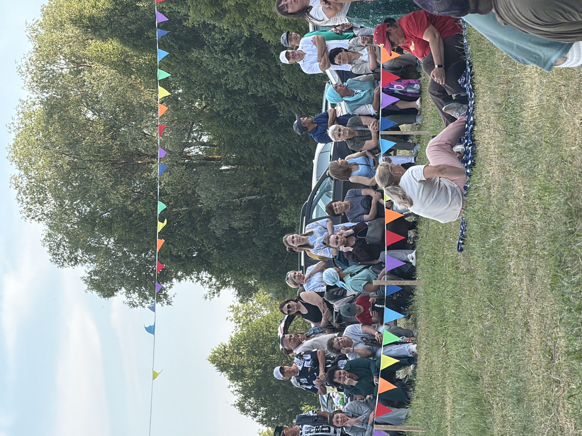In the city of Unknown, Unknown, a group of people are gathered outdoors on a grassy field, with trees and a cloudy sky visible in the background. A string of colorful triangular flags is stretched across the scene. In the foreground, two women are sitting on a blanket on the grass, facing each other. One woman, wearing a light-colored shirt and pink pants, is sitting with her legs crossed and her hands in her lap. The other woman, wearing a red shirt and dark pants, is sitting with her legs outstretched and her hands clasped. Behind them, a row of people are seated on benches, watching an event. The people are of various ages and are dressed in casual clothing. Cars are parked in the background, suggesting an event or gathering.