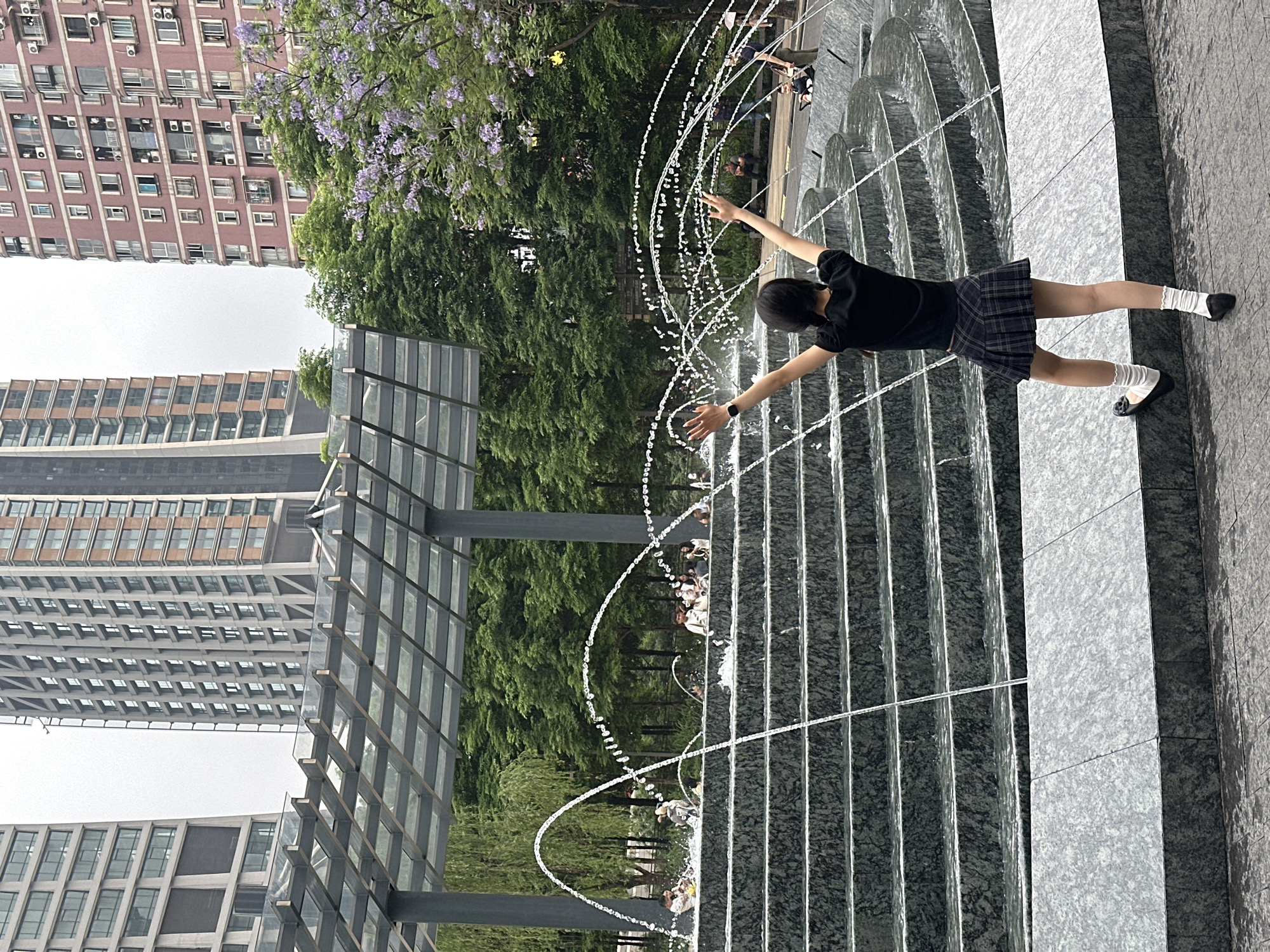 A young person stands on a textured grey stone surface, likely a walkway or platform adjacent to a water feature consisting of multiple streams of water forming arcs. The person is seen from the back, wearing a dark short-sleeved top, a plaid skirt, white socks, and black shoes. Their arms are outstretched, appearing to interact with the water streams. Behind the water feature and the person is lush greenery with hints of purple flowers, possibly jacaranda trees. In the background, tall urban buildings are visible against an overcast sky. A grey metal structure with a grid-like design is situated to the left, adding to the urban architectural landscape. The location is likely in Yuzhong District, China, given the urban architecture and the presence of specific flora.