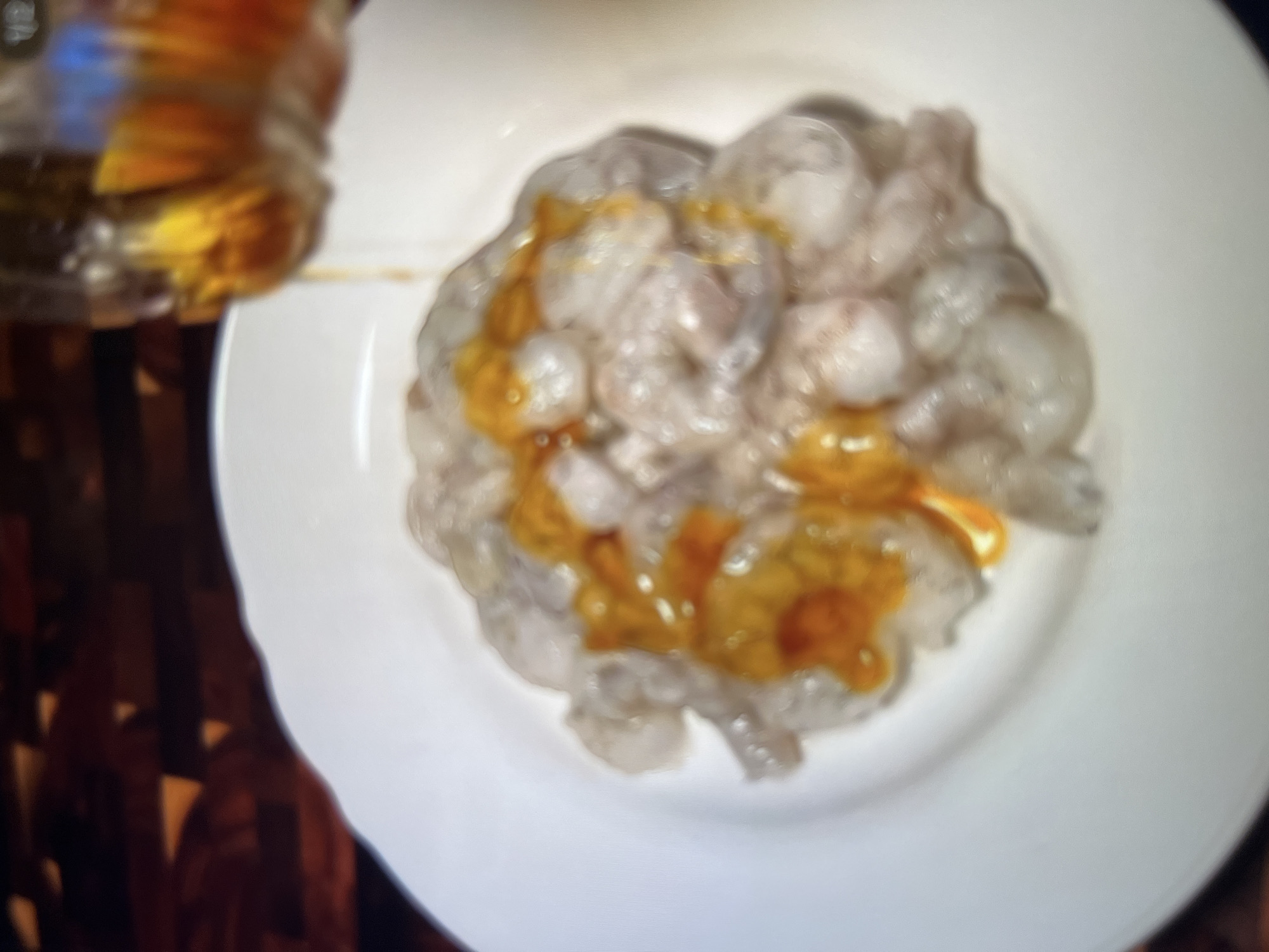 In Abuja, Nigeria, a close-up shot captures a white plate filled with raw shrimp. A stream of amber liquid, likely oil or a sauce, is being poured from a blurred bottle, drizzling over the shrimp. The background features a dark, textured surface, possibly a wooden cutting board. The lighting is dim, with some reflections on the plate and bottle, creating a culinary scene.