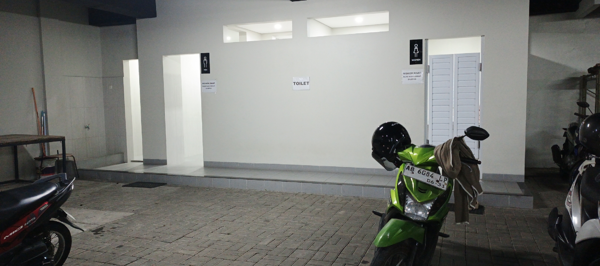 The image depicts an outdoor scene at night, likely a public restroom facility located in Godean, Indonesia, given the license plate AB 6084 LP which is typical for Yogyakarta province. In the foreground, a bright green scooter is parked, with a black helmet resting on its handlebars and a brown jacket draped over the handlebars. The scooter's license plate is clearly visible. To the left of the scooter, a portion of a red and black Yamaha scooter is also visible. The background features a building with distinct sections for men's and women's restrooms, indicated by signage. The entrance to the men's restroom shows an open doorway leading into a brightly lit interior, with a sign that reads "MOHON MAAF ALAS KAKI HARAP DILEPAS" which translates to "SORRY, SHOES MUST BE REMOVED." The women's restroom has closed louvered doors. A sign that reads "TOILET" is also present on the wall. The ground is paved with interlocking stone tiles. In the far left of the image, a metal table and a bucket are visible. To the right, other parked motorcycles are partially visible. The overall lighting suggests it is evening or night, with artificial lights illuminating the area.