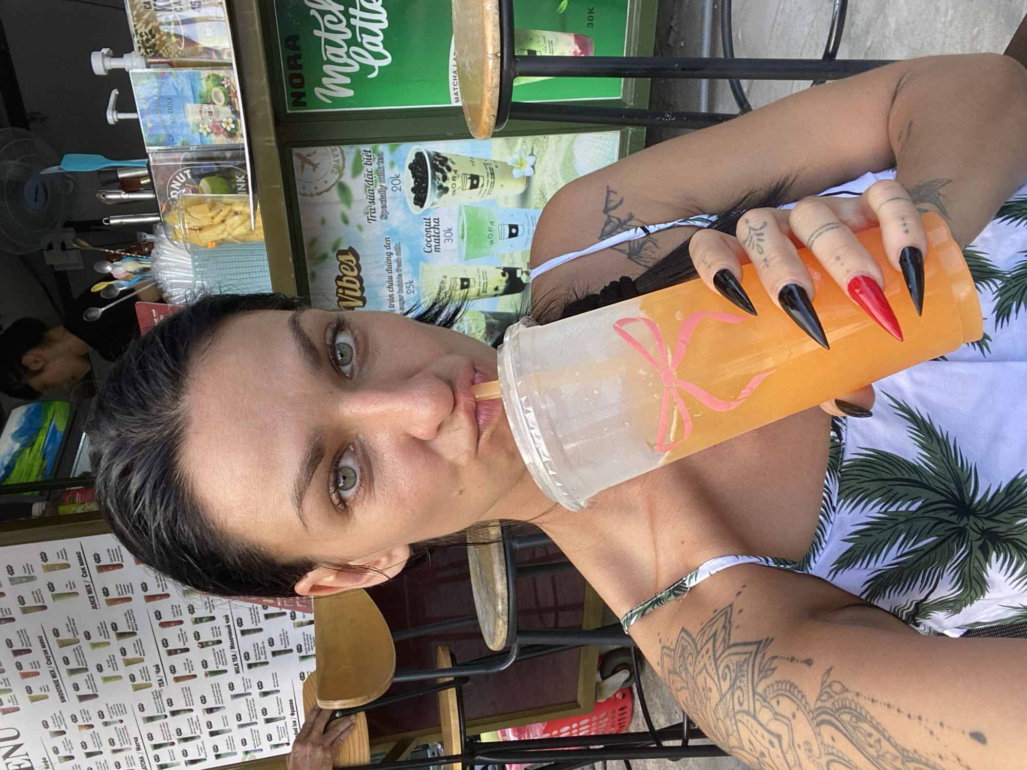 A woman with fair skin and dark hair is shown in a selfie while drinking from a large plastic cup with a straw. She is wearing a white swimsuit with a green palm tree pattern. Her nails are long and pointed, colored black and red. The woman has a tattoo on her arm. The photo was taken outdoors in Phu Thuy Ward, Vietnam, based on the menu visible in the background. The menu features drinks with prices listed in Vietnamese currency. The area around the woman appears to be a cafe or a street-side vendor, with a table, chairs, and a menu display.